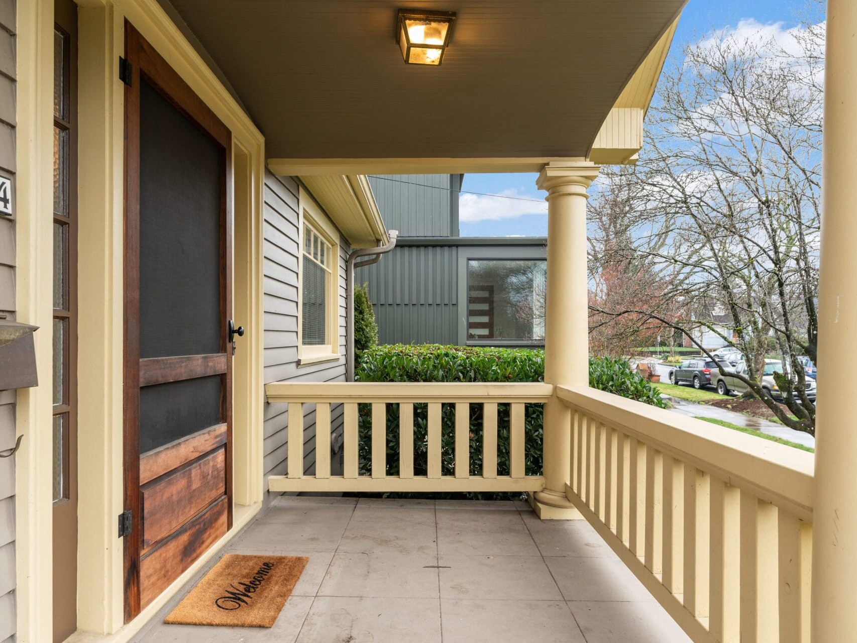 A front porch with beige railings and columns, featuring a wooden door and a welcome mat. The siding is gray, and theres a small mailbox on the left. Trees and parked cars are visible in the background under a partly cloudy sky.