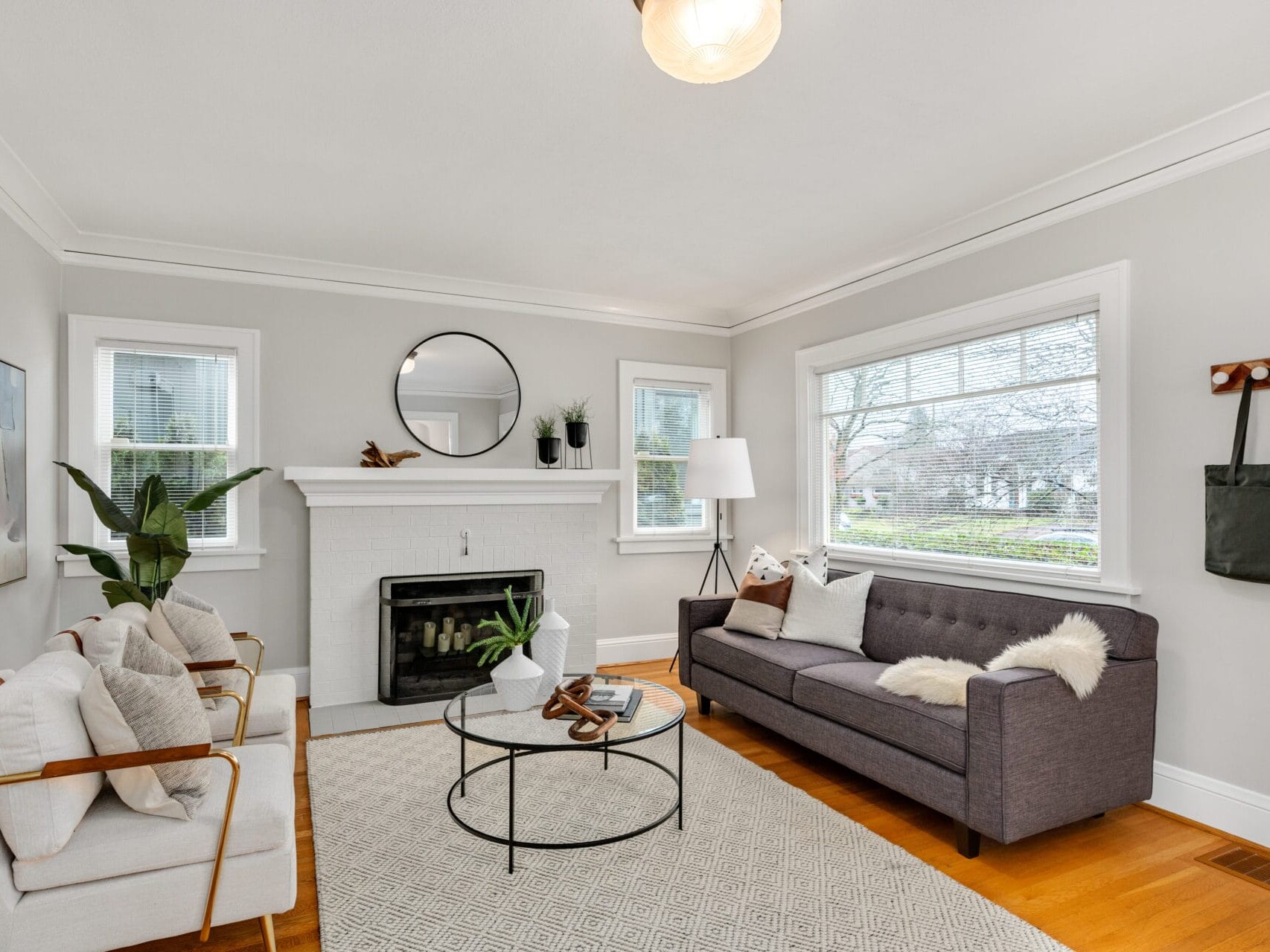 A cozy living room with light gray walls features a dark gray sofa, two white armchairs, and a round glass coffee table. A white brick fireplace is adorned with a round mirror and plants, and a large window lets in natural light.