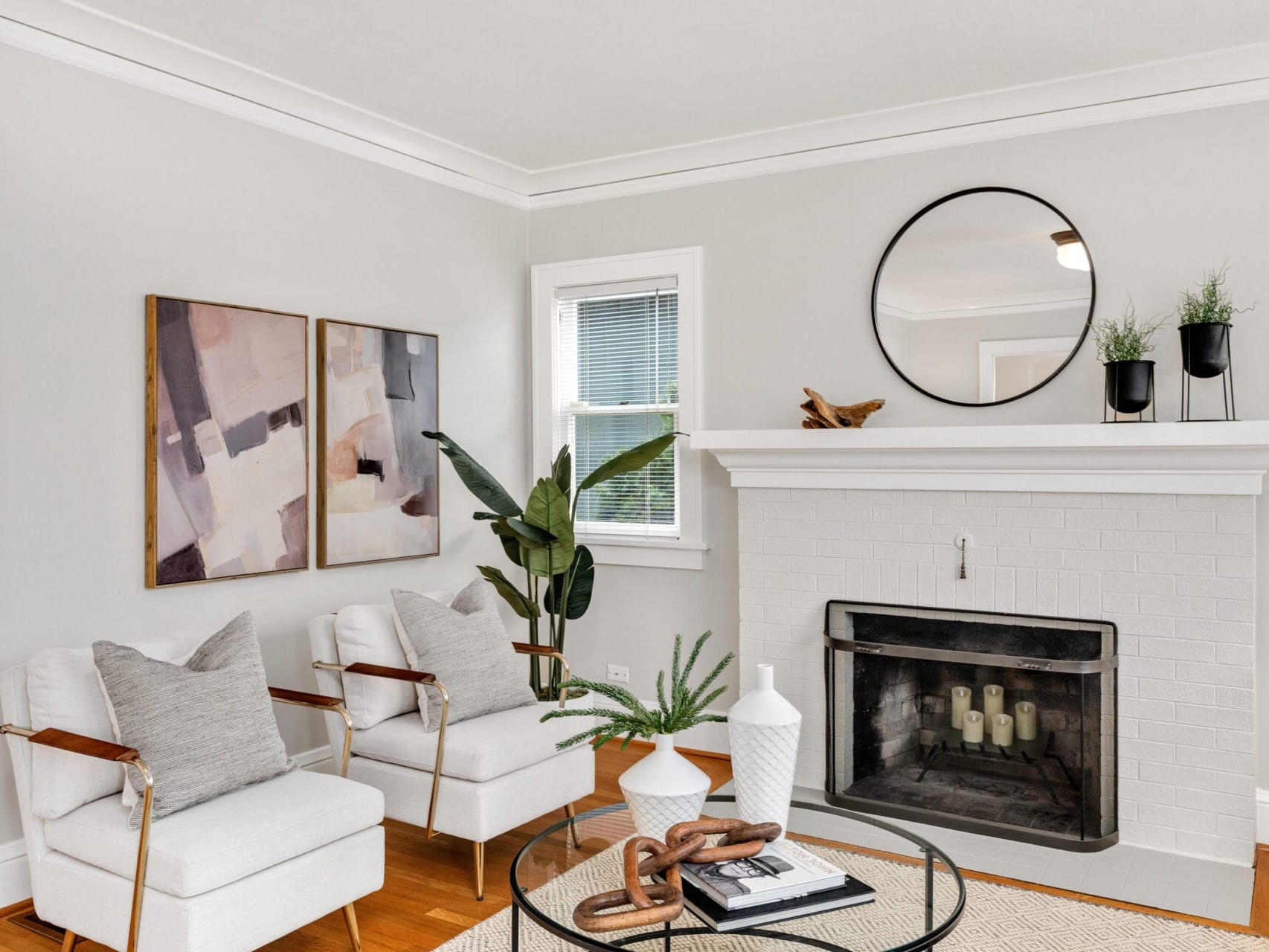 A cozy living room with two white armchairs and gray pillows near a brick fireplace. A round mirror, plants, and abstract art decorate the walls. A small coffee table with vases and books sits on a woven rug, enhancing the rooms neutral tones.