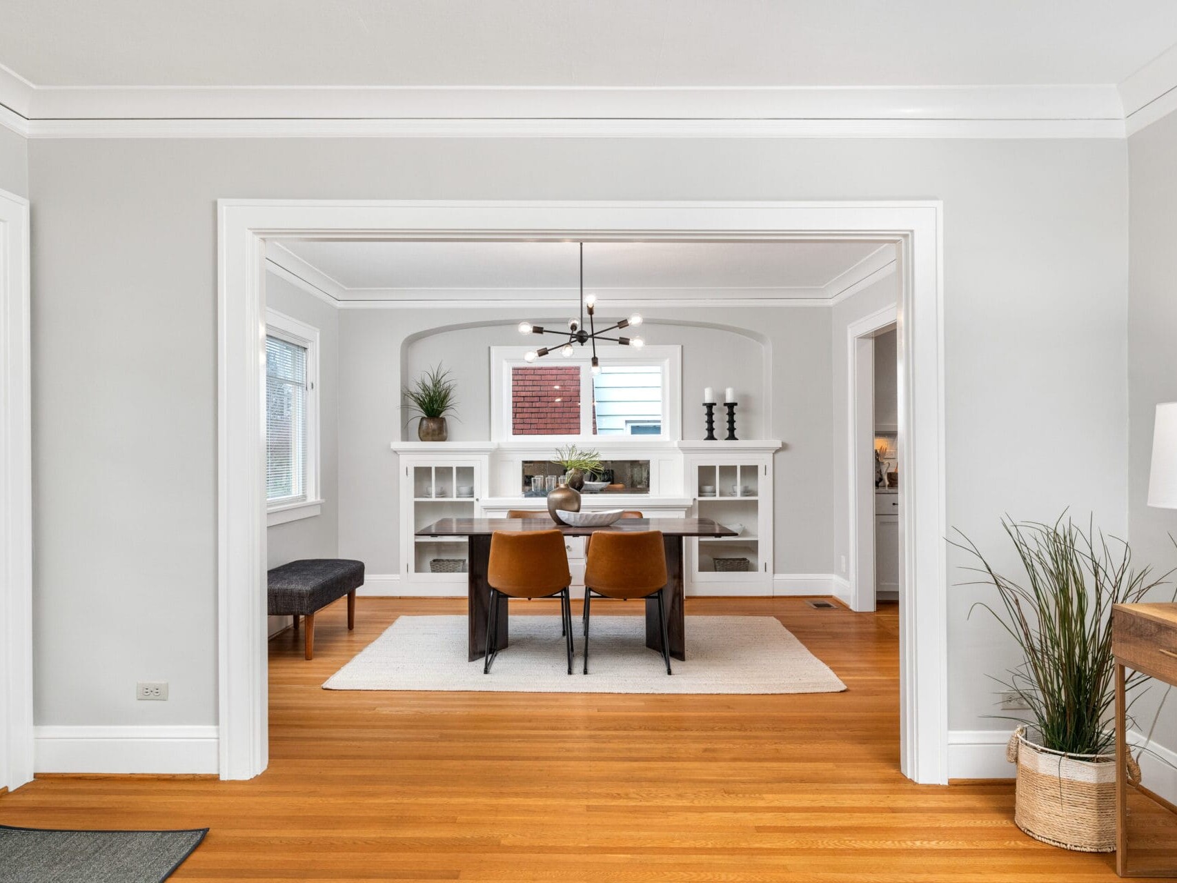 A modern dining room with a wooden floor and a white rug. It features a table with four brown chairs, a contemporary chandelier, built-in shelving with decorative items, and natural light from the windows. A plant is placed in a pot on the right.