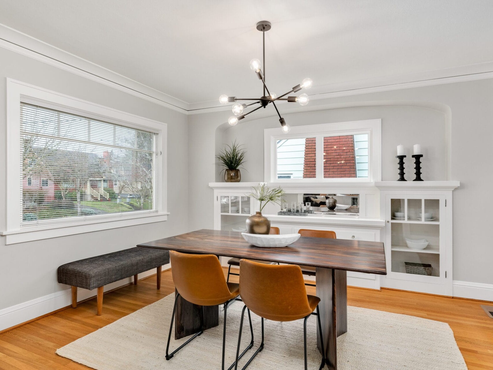 A dining room with a modern wooden table and two brown chairs. A bench lines one wall under a large window, while a white sideboard is decorated with plants and candles. A contemporary chandelier hangs above the table on a wooden floor.