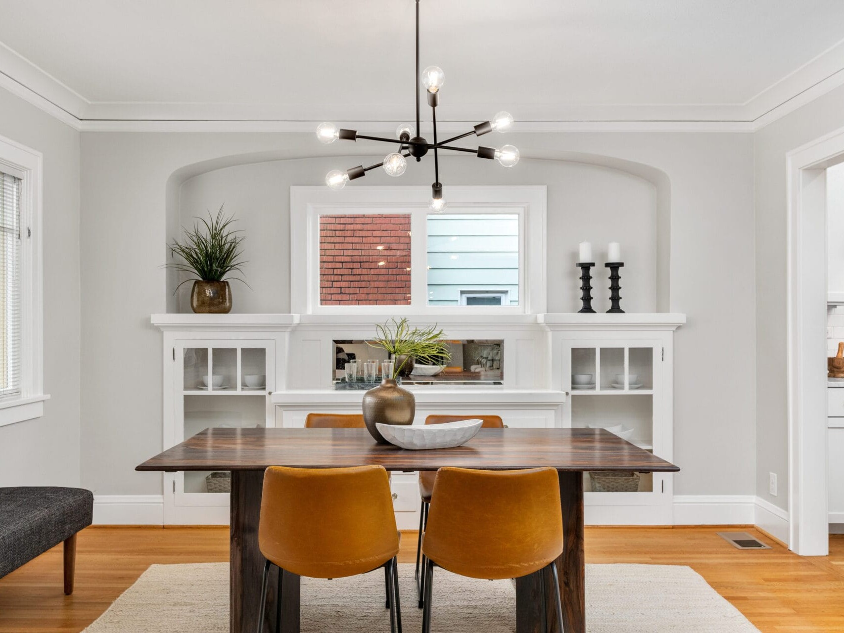 A modern dining room with a wooden table and four mustard-colored chairs. A chandelier with exposed bulbs hangs above. Built-in shelves hold plants and candles. The room has white walls, a window, and a doorway to a kitchen in the background.