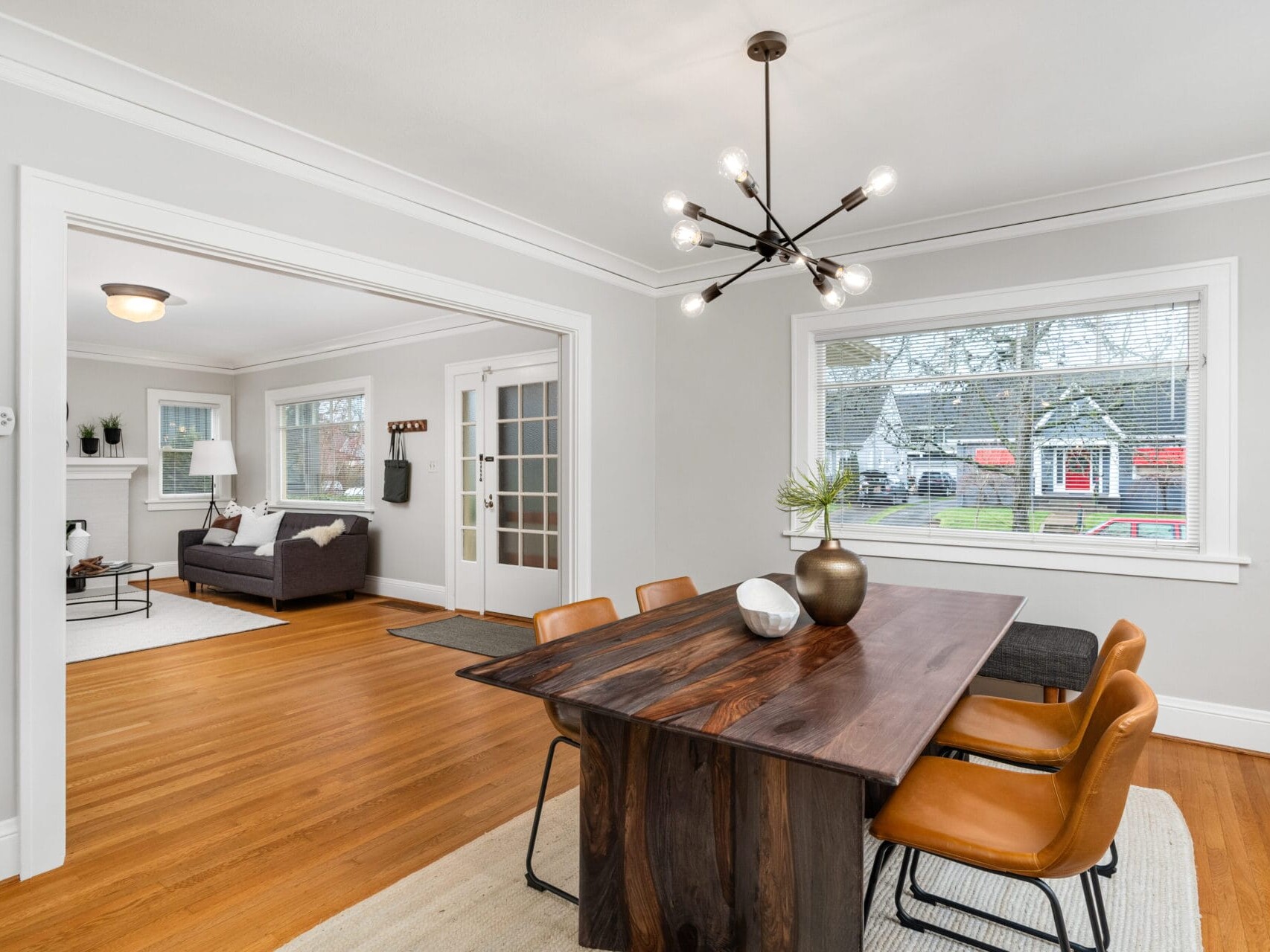 A modern dining room with a wooden table and four orange chairs. A unique chandelier hangs above. The room has hardwood floors and a large window showing the outdoors. A cozy living area is visible in the background.