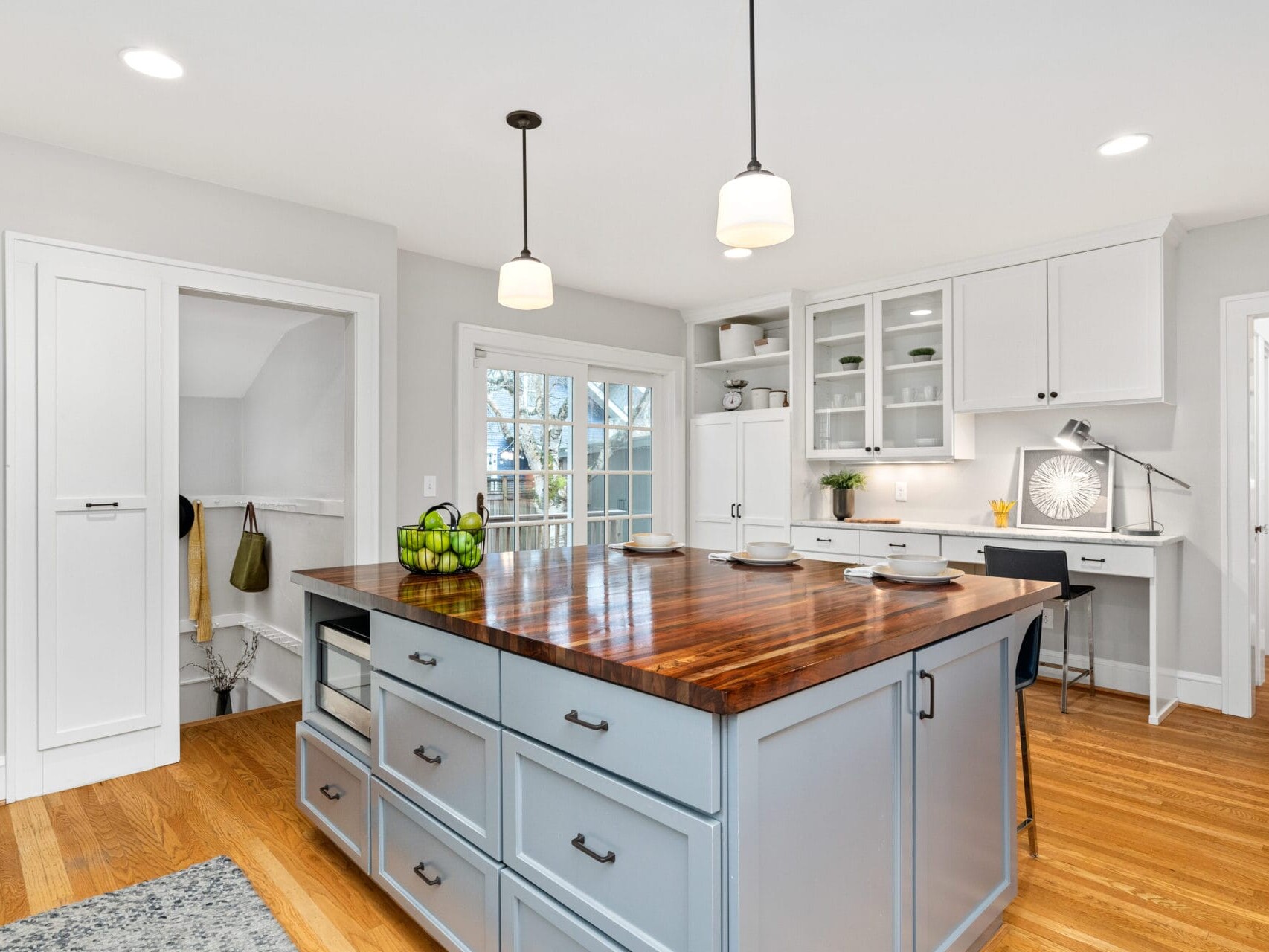 A modern kitchen with light gray cabinets and a wooden island countertop. The kitchen features pendant lights, glass cabinet doors, and French doors leading outside. There are stools around the island, and a bowl of green apples on the counter.