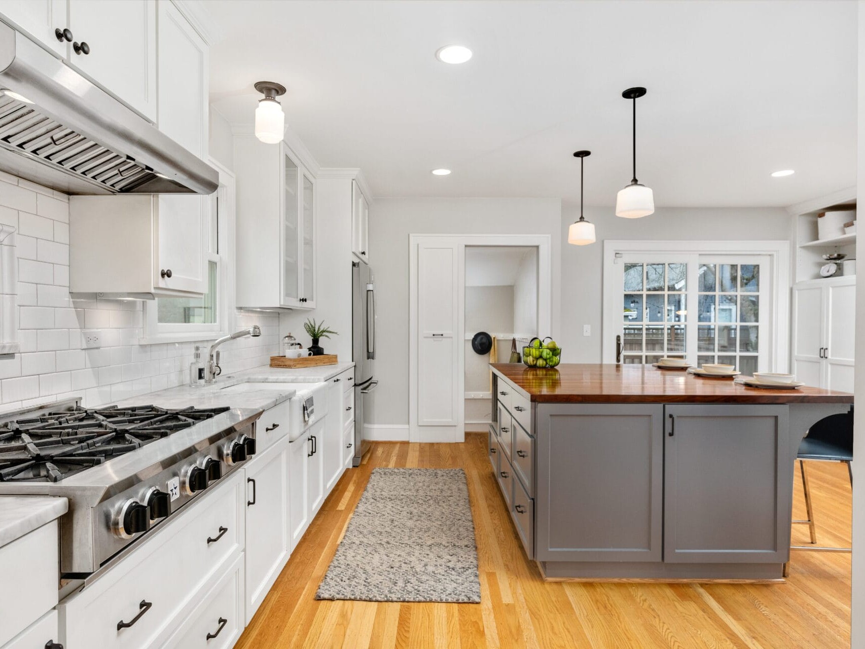 Spacious modern kitchen with white cabinetry, stainless steel appliances, and a large gray island with pendant lights. Hardwood floors complement the bright, airy space, which includes a dining area with large windows.