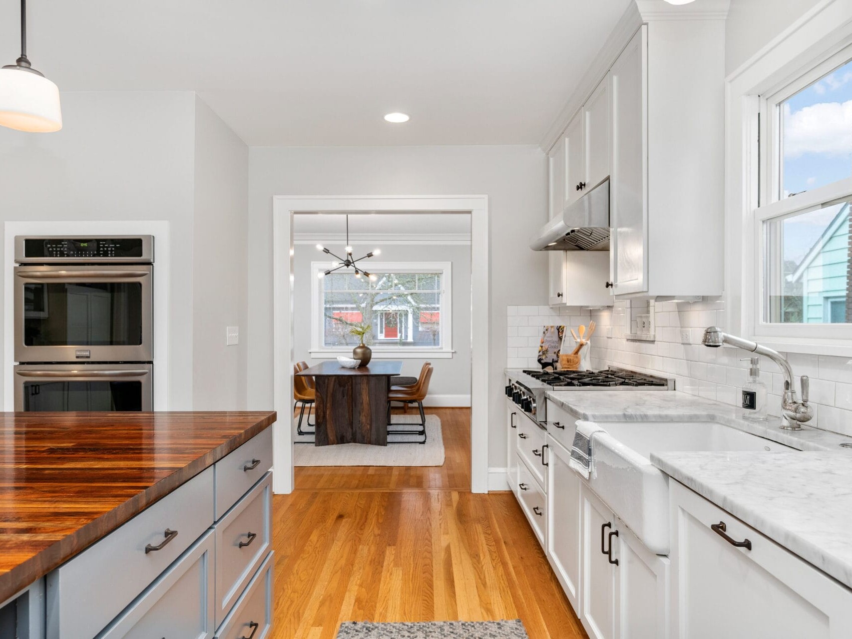 A bright kitchen with white cabinetry, marble countertops, and stainless steel appliances. A wooden island is in the foreground. Beyond, a dining area with a wooden table and chairs is visible through a wide doorway. Wood floors add warmth.