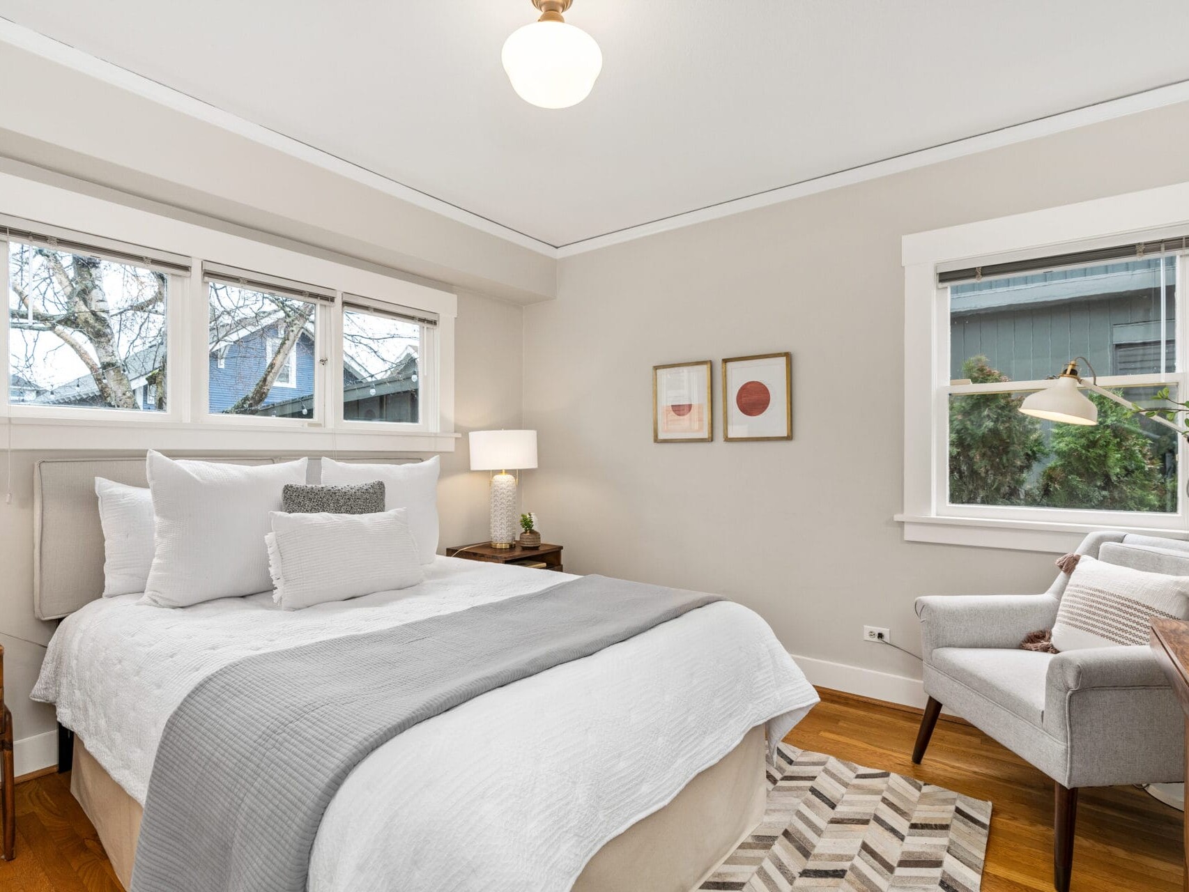 A cozy bedroom with a neatly made bed featuring white and gray linens. The room includes two nightstands with lamps, a gray armchair, a plant, and framed artwork on the walls. Natural light streams in through the windows.