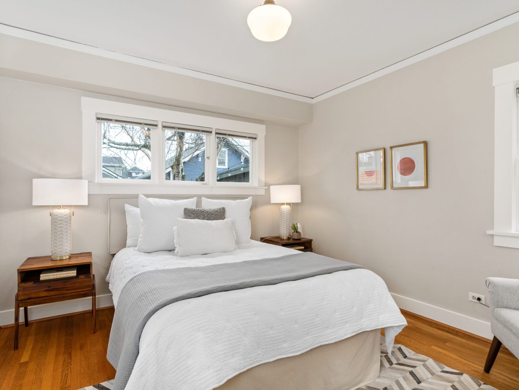 A cozy bedroom with a neatly made bed featuring white and gray bedding. Two wooden nightstands flank the bed, each with a lamp. A beige armchair sits to the right, and three framed artworks are on the walls. A window offers a view outside.