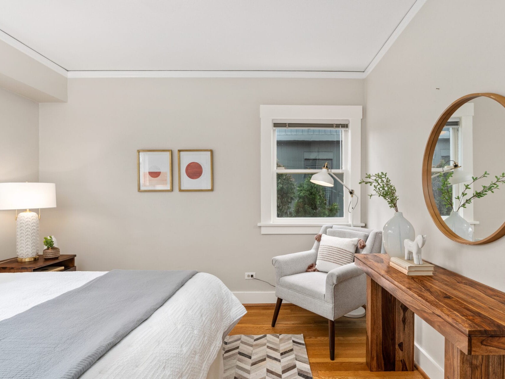 A cozy bedroom features a neatly made bed with white and gray bedding. The room includes a wooden console table, a round mirror, a gray armchair, and two framed artworks. A window lets in natural light, illuminating the minimalist decor.