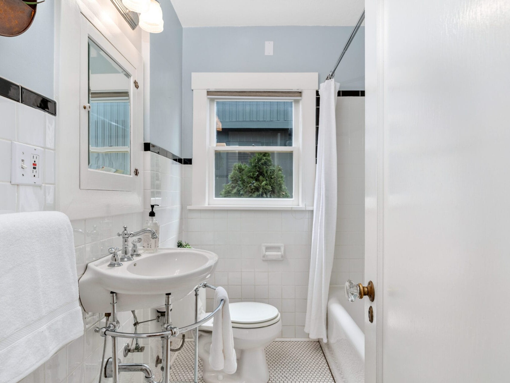 A small, bright bathroom with light blue walls and white tiles. It features a white sink, toilet, and bathtub with a shower curtain. A window above the toilet shows an outdoor view. Towels hang on the wall, and a mirror is above the sink.