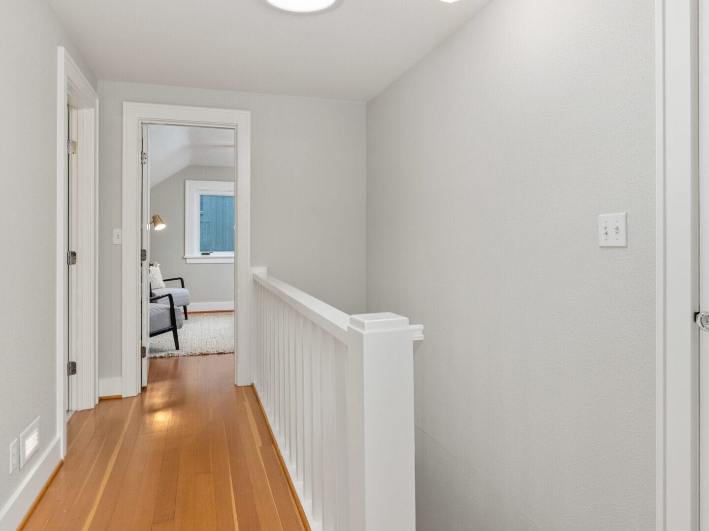 A bright hallway with wooden floors and white walls. A handrail runs along one side, leading to a room with a bed and chair visible in the distance. The ceiling features a light fixture and a round skylight, while modern art hangs on the wall.