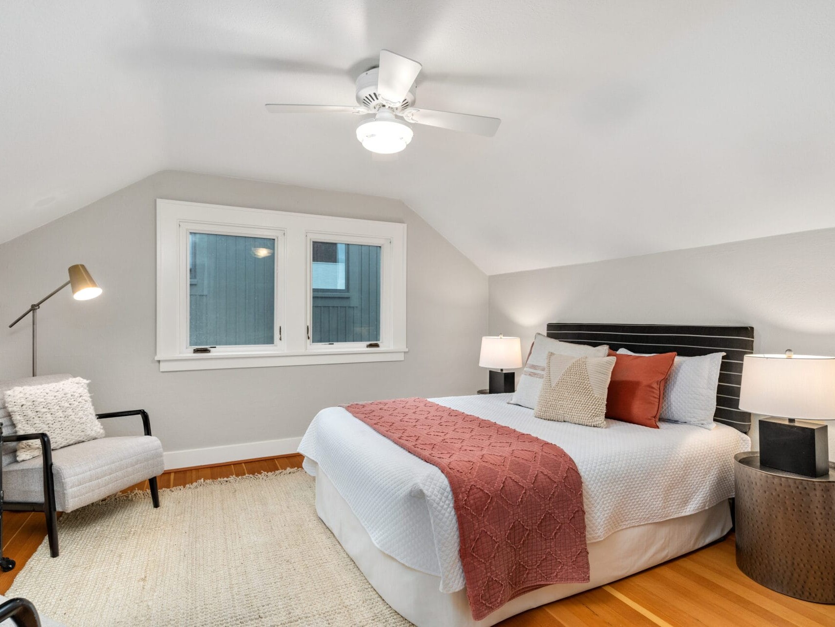 A cozy bedroom with a neatly made bed featuring white and pink bedding. Two bedside tables with lamps flank the bed. Theres a gray armchair with a floor lamp beside it. A ceiling fan hangs above and a window lets in natural light. A rug covers the wooden floor.