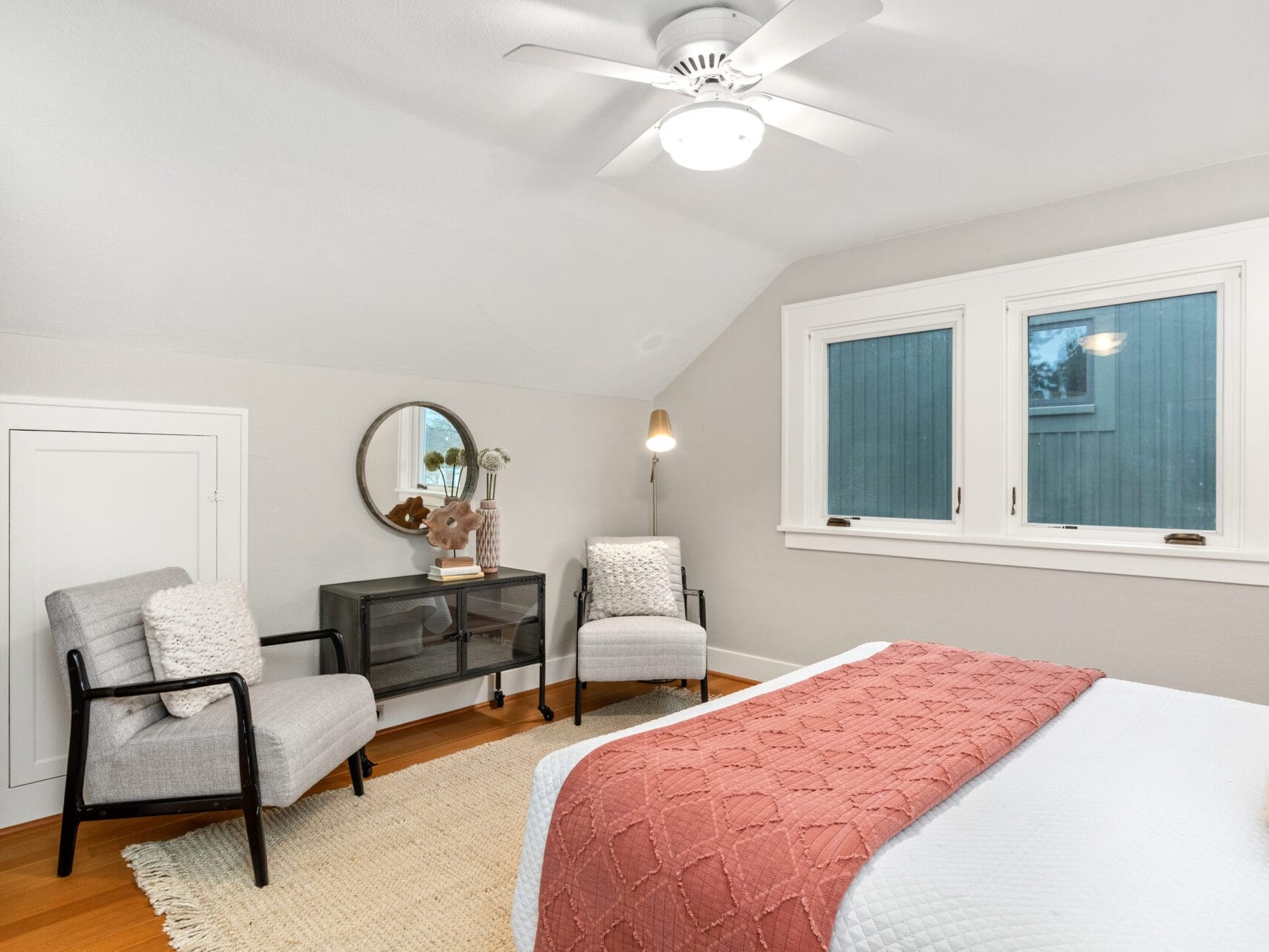 A cozy bedroom with a bed featuring a quilted coral blanket. Two gray chairs sit around a small glass table under a circular mirror. The room has a light tan wall, a ceiling fan, and two windows, with hardwood floors and a beige rug.