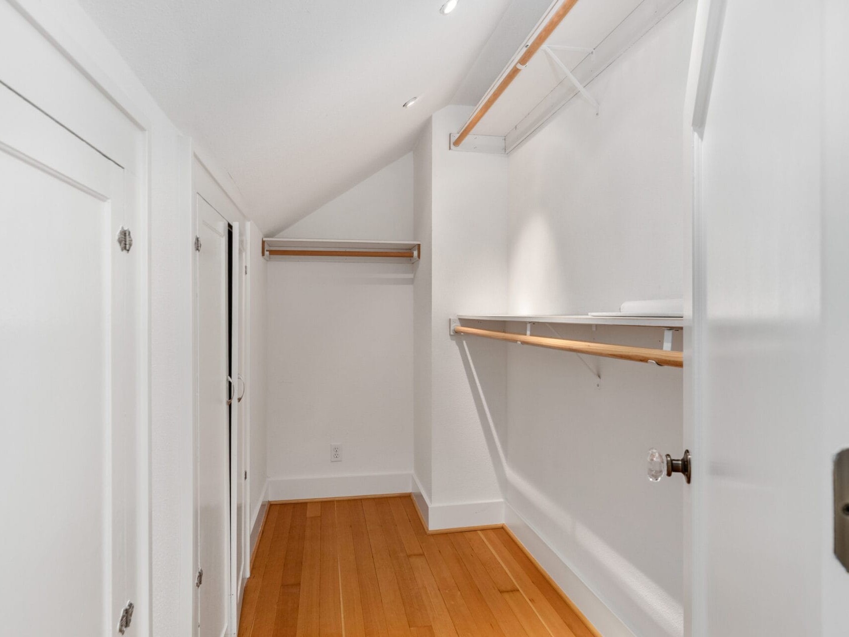 A narrow, white walk-in closet with wooden shelves and hanging rods on both sides. The floor is made of light-colored wood, and the ceiling is slanted. The space is well-lit with recessed lighting.