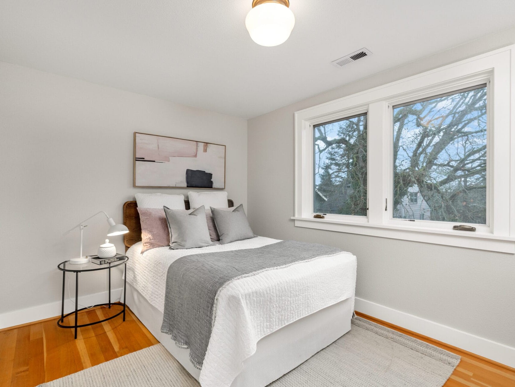 A cozy bedroom with a neatly made bed featuring white and gray bedding. A small round bedside table with two white lamps is beside the bed. A large window provides natural light, and a modern abstract painting hangs above the bed.