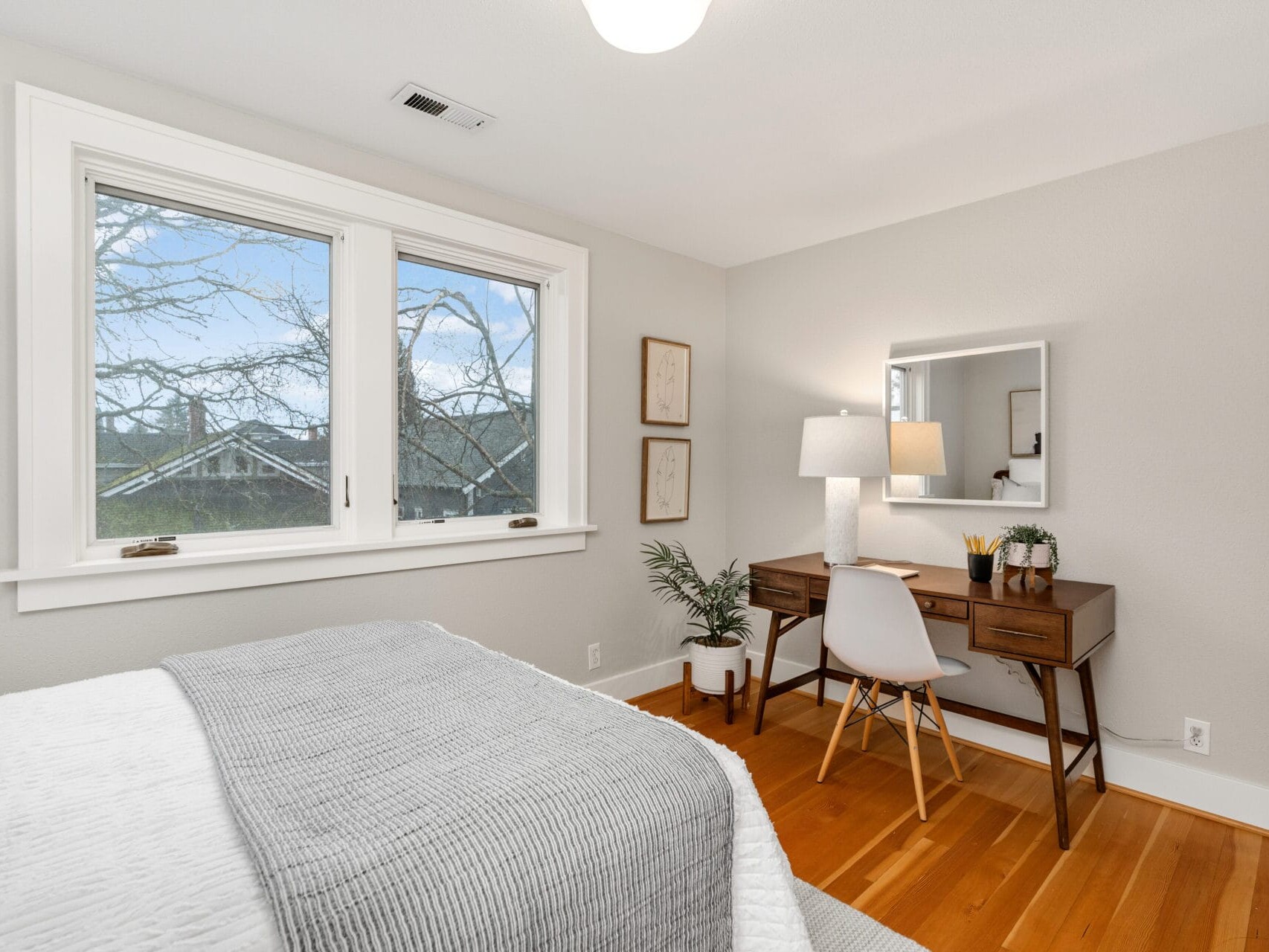 A tidy bedroom with a bed covered in white and gray bedding. A wooden desk with a mirror and lamp is in the corner, accompanied by a white chair. A plant decorates the desk area, and a window shows a view of trees and a house.