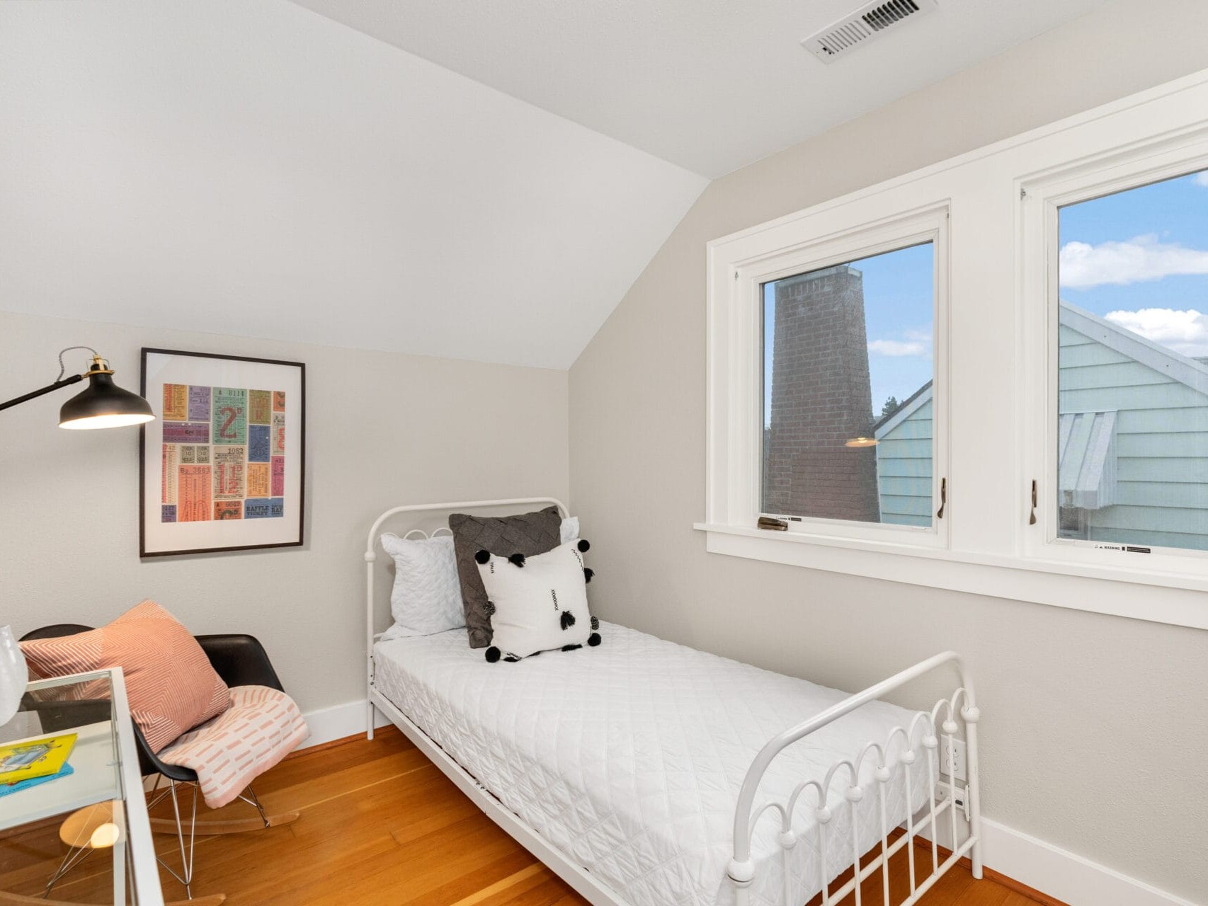 A small bedroom with a white metal bedframe and white bedding. Theres a panda pillow on the bed. A black chair with a pink cushion sits beside a desk with a lamp. A colorful framed artwork hangs on the wall. Large windows let in natural light.