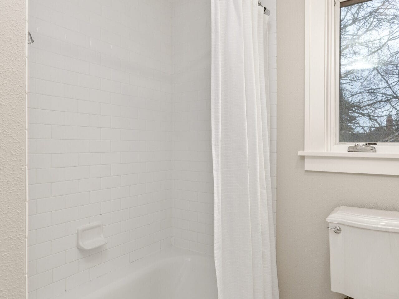 A bathroom features a white bathtub with a shower curtain, white subway tile walls, and a small built-in shelf. There is a white toilet next to a window with tree branches visible outside. The floor has white hexagonal tiles.