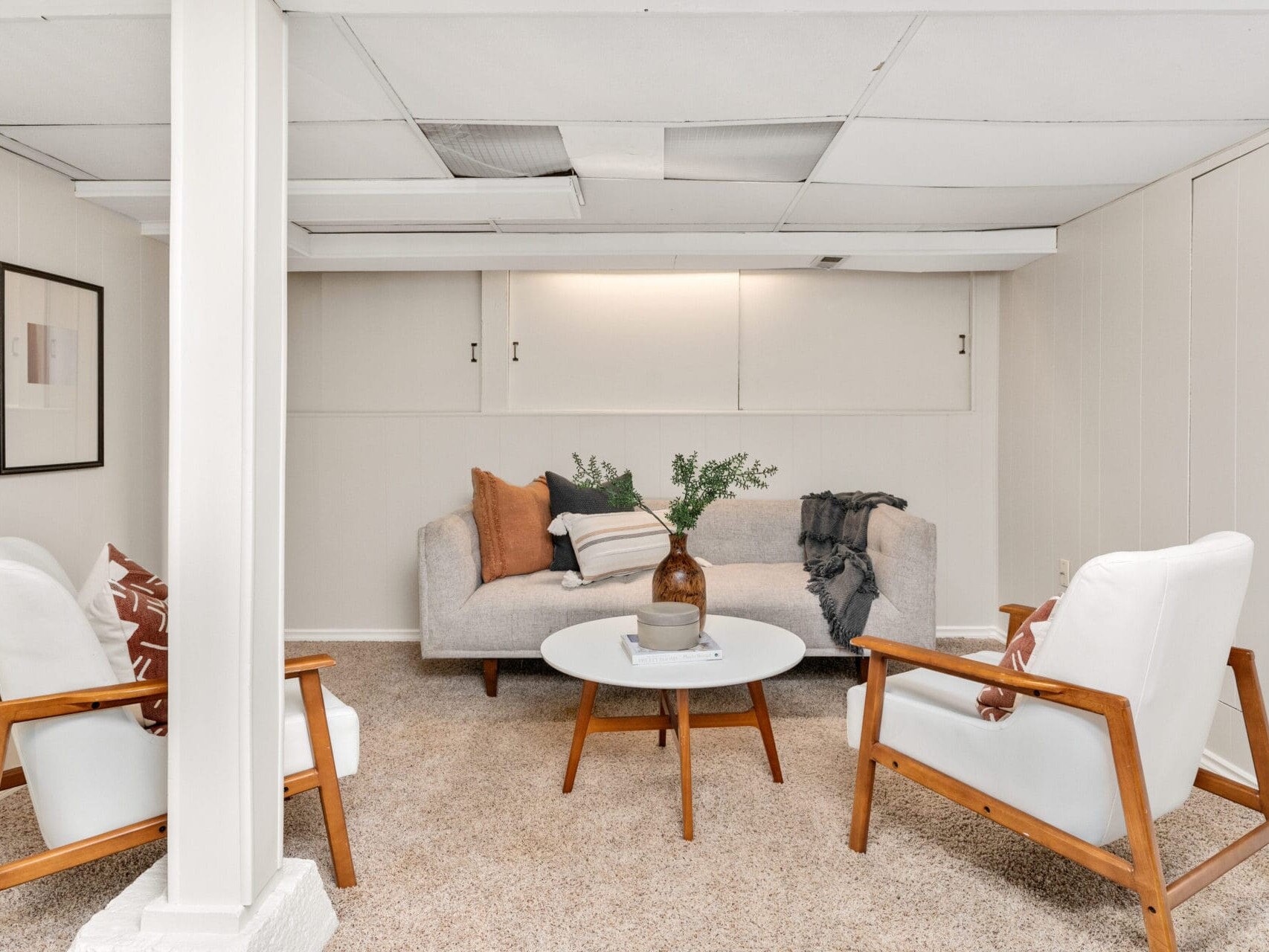 A cozy living room with a beige sofa and two wooden armchairs with white cushions arranged around a circular coffee table. The room features neutral walls, framed artwork, and a soft carpet. A vase with greenery decorates the table.