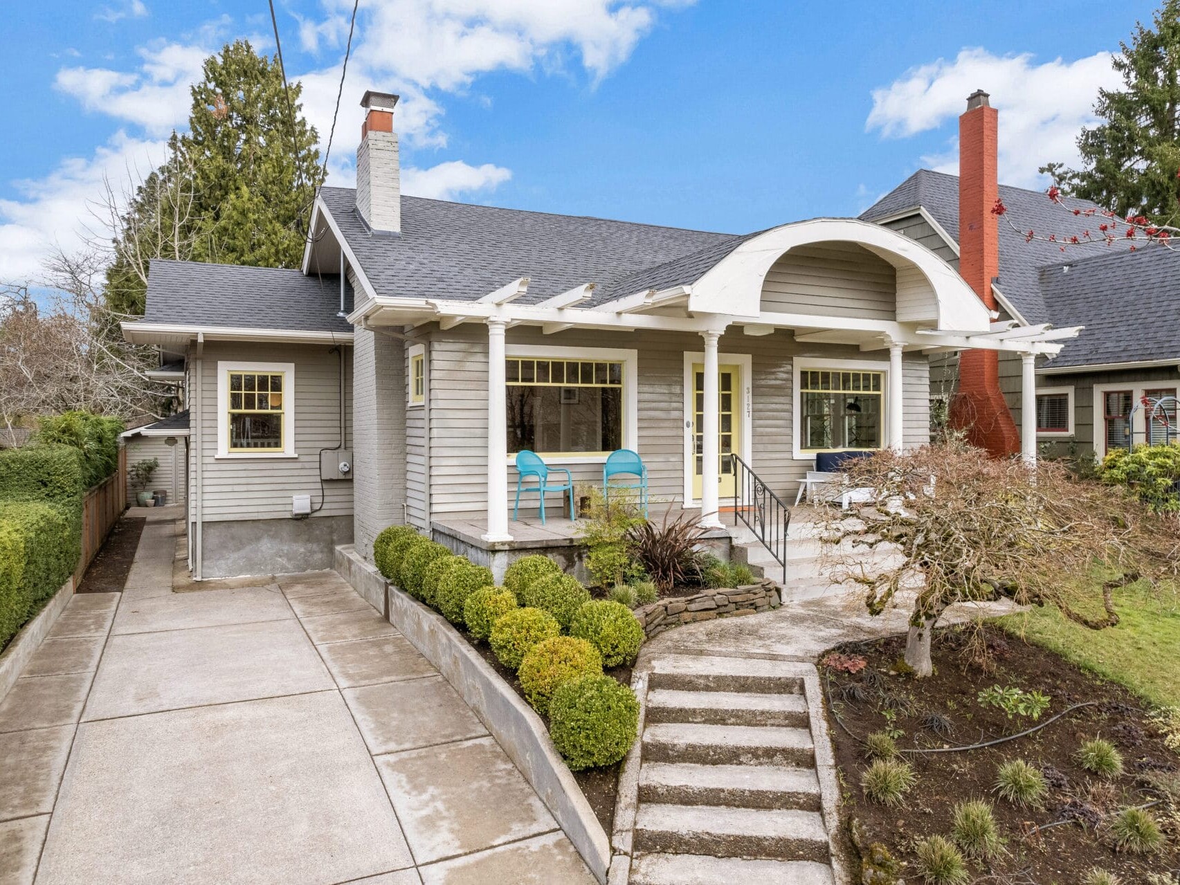 Charming gray bungalow with a sheltered front porch, two blue chairs, and a landscaped front yard. A pathway leads to the entrance, and a driveway extends to the side. The sky is clear and blue.
