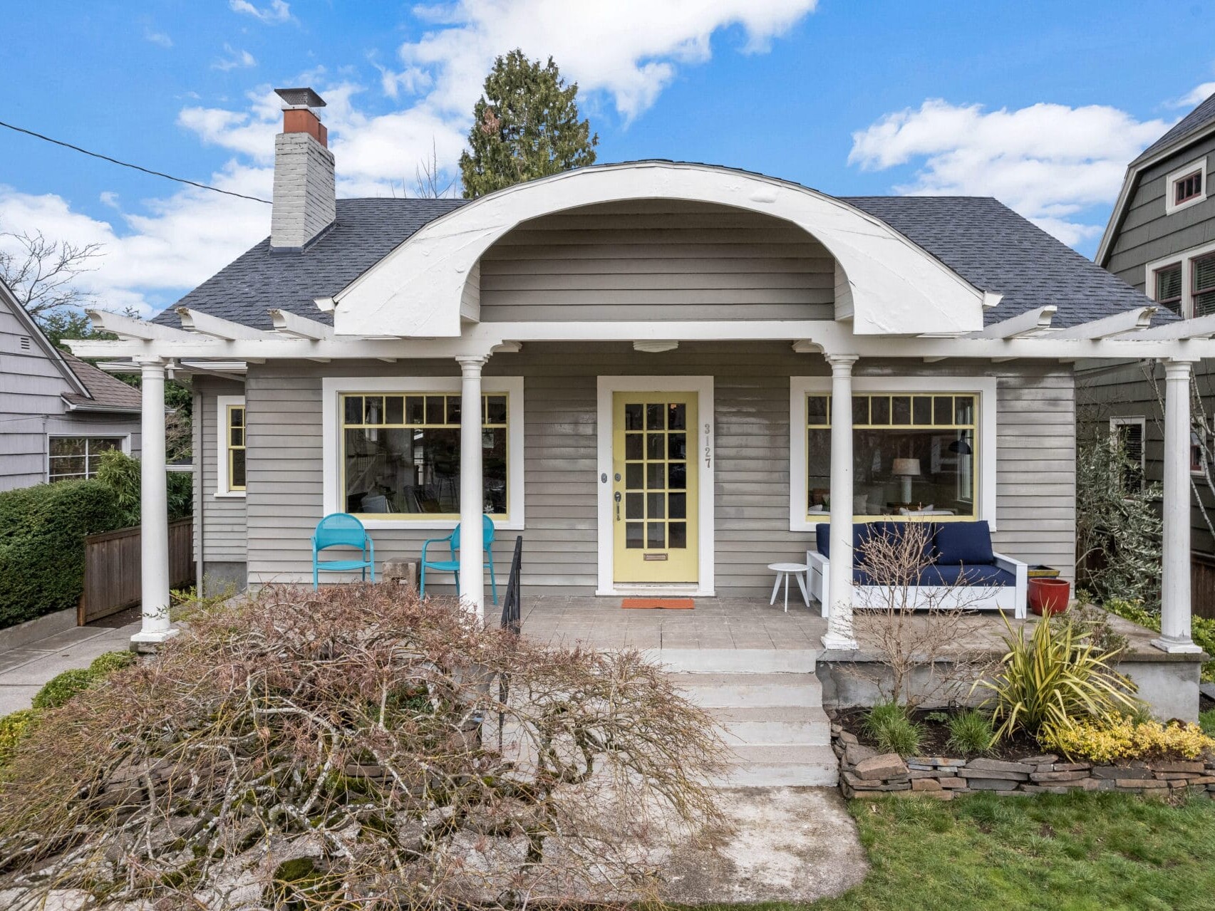 A charming gray house with a yellow door, white trim, and a large front porch. The porch features two chairs and a small sofa. There are bushes and a small tree in the landscaped front yard. The roof is dark gray with a chimney. Blue sky in the background.