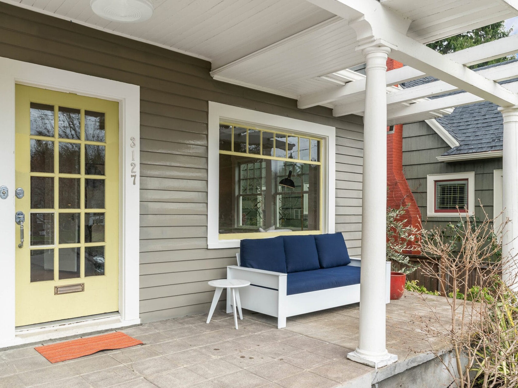 A cozy front porch with a pale yellow door and large window on a gray house. A white bench with blue cushions and a small round white table sit on the tiled porch floor. White columns support the overhead structure. Red brick and greenery are visible.