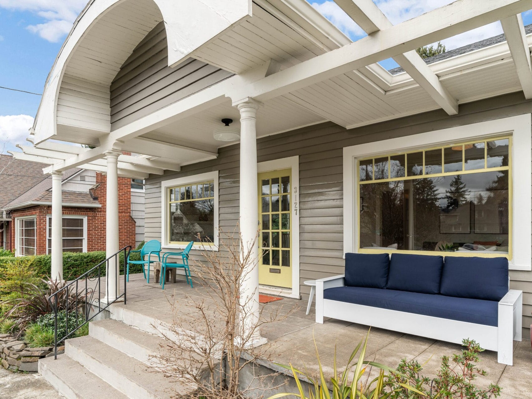 A cozy gray house features a welcoming front porch with blue outdoor furniture, including a sofa and chairs. Lush greenery and a brick house are in the background, enhancing the inviting and serene atmosphere.