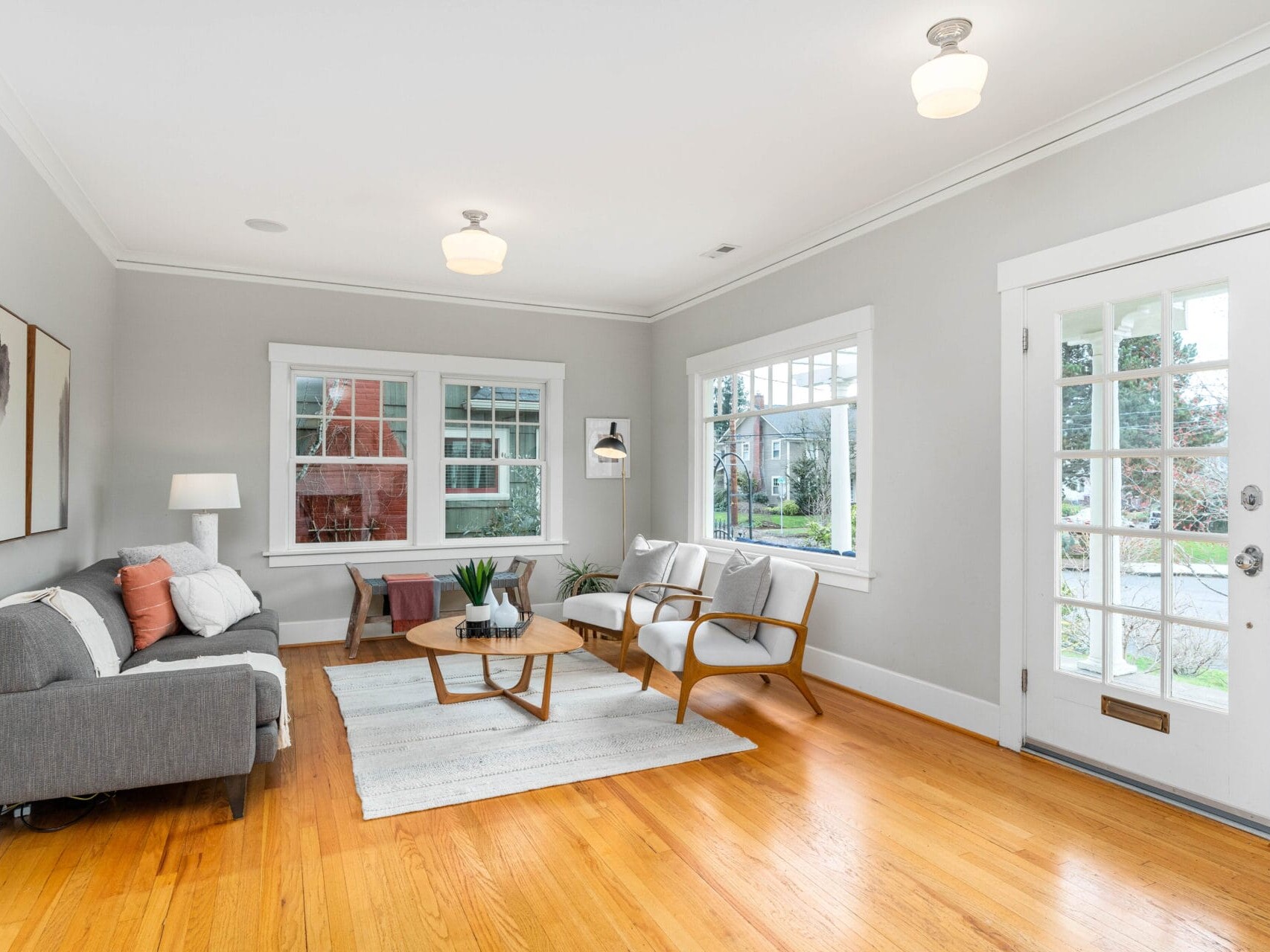 A bright, modern living room with a gray sofa, wooden coffee table, and two armchairs. Large windows let in natural light. A rug covers the hardwood floor, and a door with glass panels leads outside. A minimalistic painting hangs on the wall.