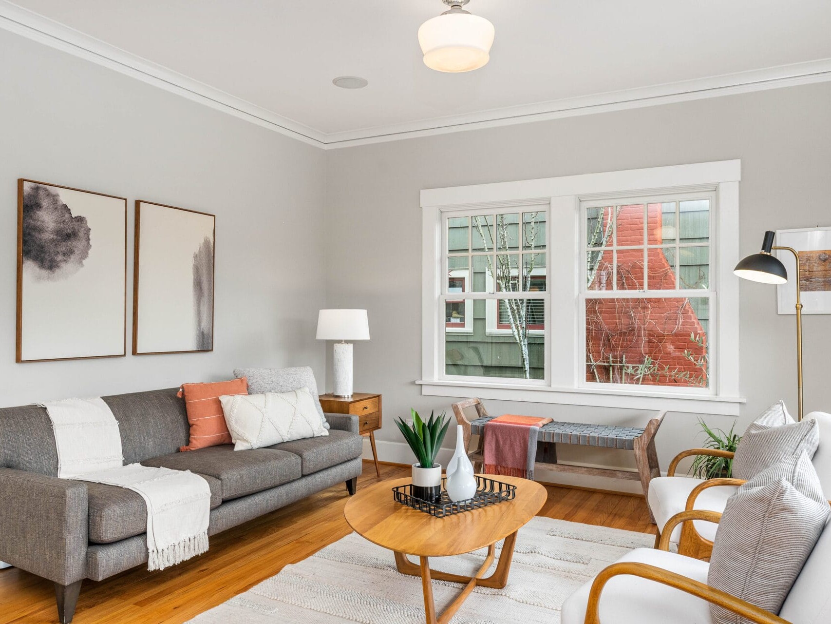 Bright living room with a gray sofa, white armchairs, and wooden coffee table. Large window, abstract art on the wall, floor lamp, and cozy decor create a welcoming atmosphere. Soft, neutral color palette with accents of orange and green.