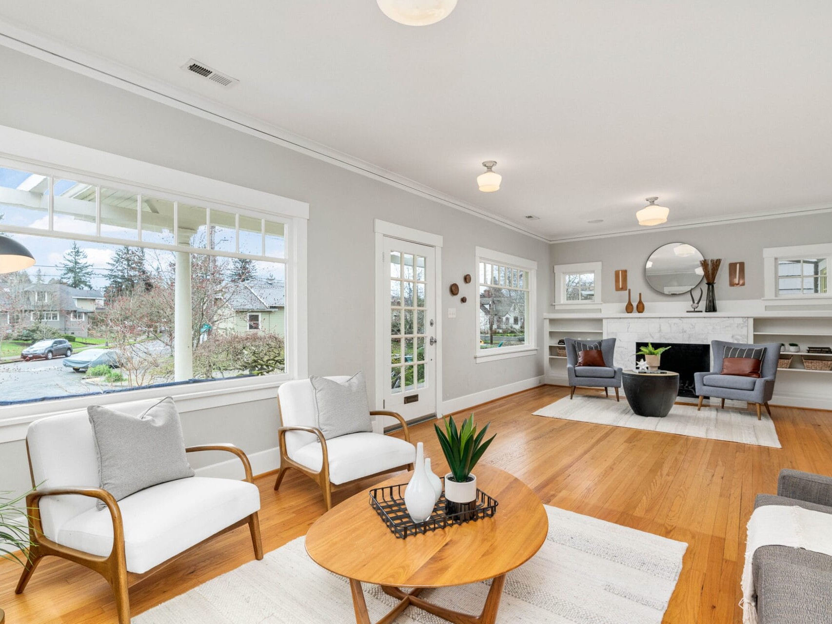 A bright, spacious living room with wooden floors and a large window. It features two white armchairs, a round wooden coffee table with a plant, a fireplace with a mirror above, and two dark gray chairs. White walls add to the airy atmosphere.