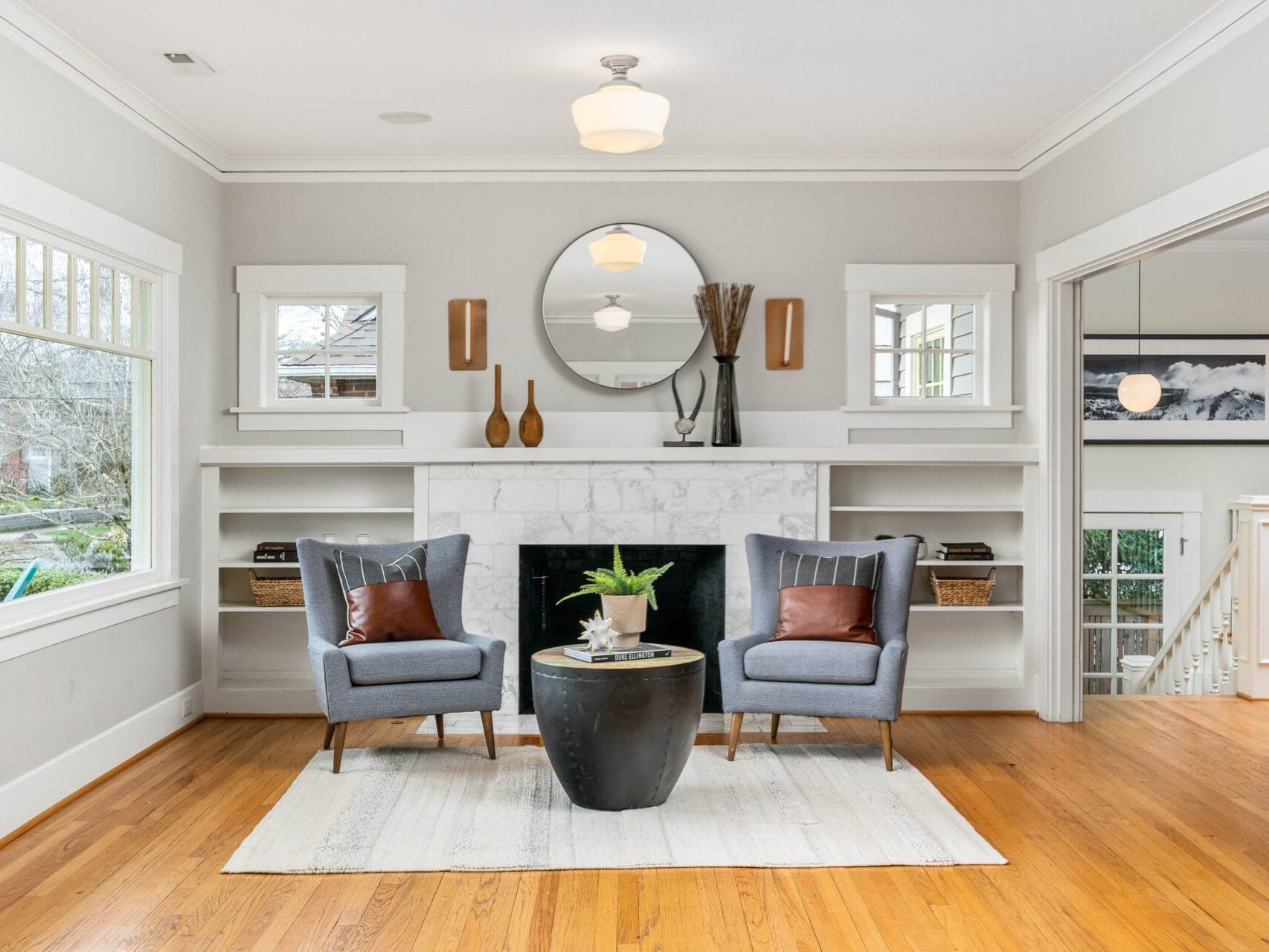 A cozy living room with two gray armchairs facing a black round table on a white rug. A white fireplace with a circular mirror above is flanked by built-in shelves. Large windows and light wood floors add brightness to the space.