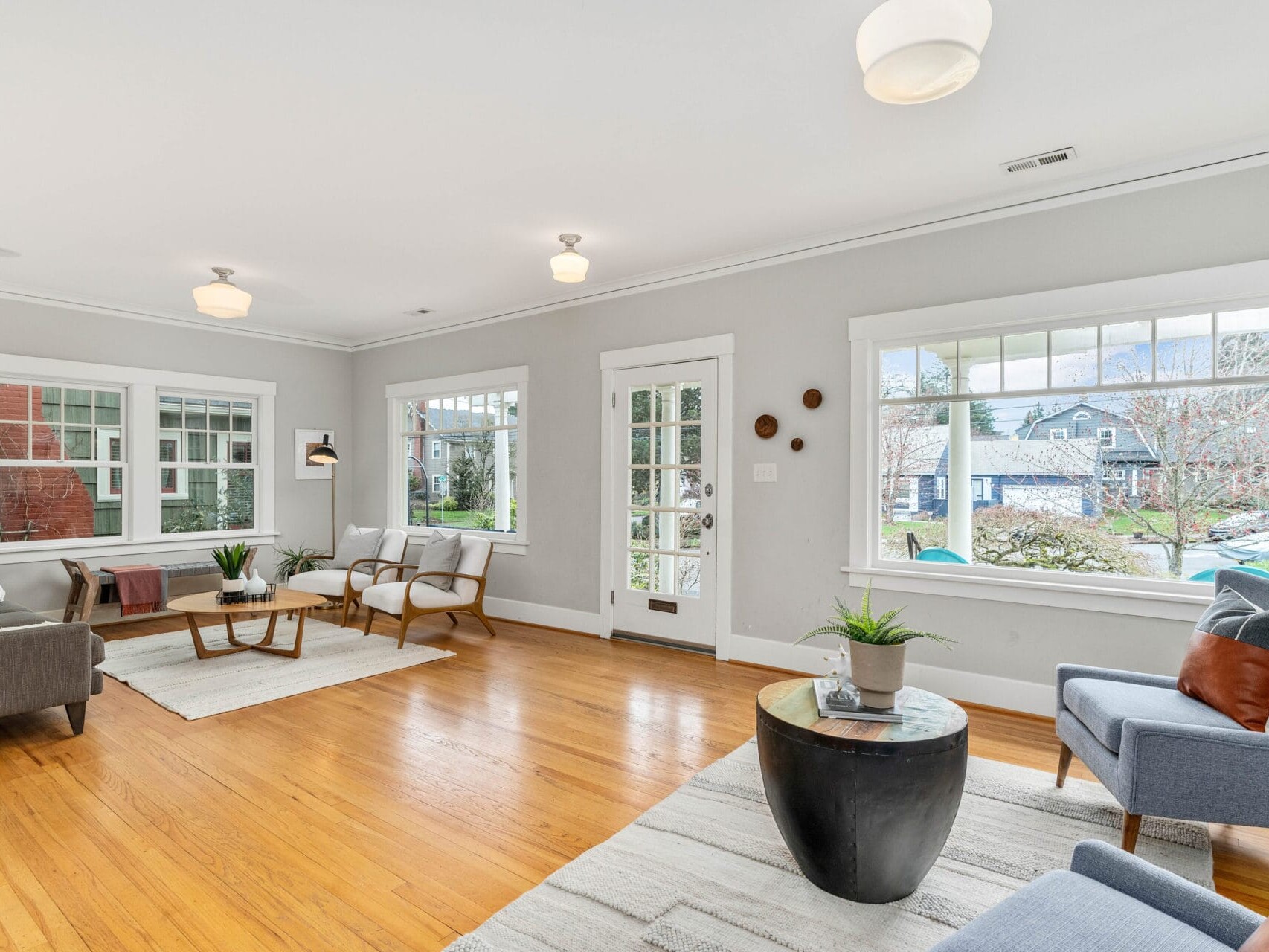 A bright living room with large windows, wooden flooring, and light gray walls. It features modern furniture including a gray sofa, chairs, a wooden coffee table, and a small round side table. A door leads to an outdoor view of trees and a house.