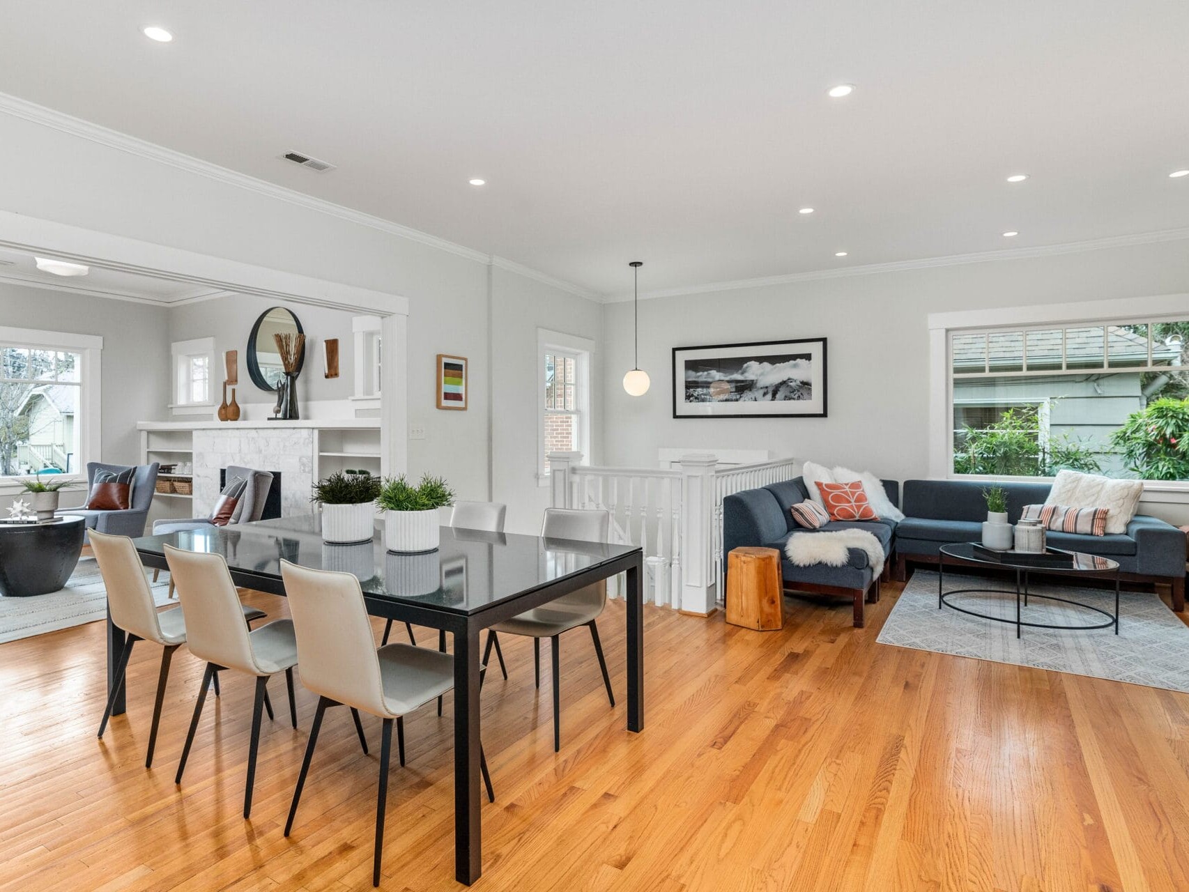 A modern living and dining area with hardwood floors. The room features a black dining table with gray chairs, a blue sofa, a white fireplace, and large windows. Decor includes plants, a mirror, and framed artwork. A pendant light hangs from the ceiling.