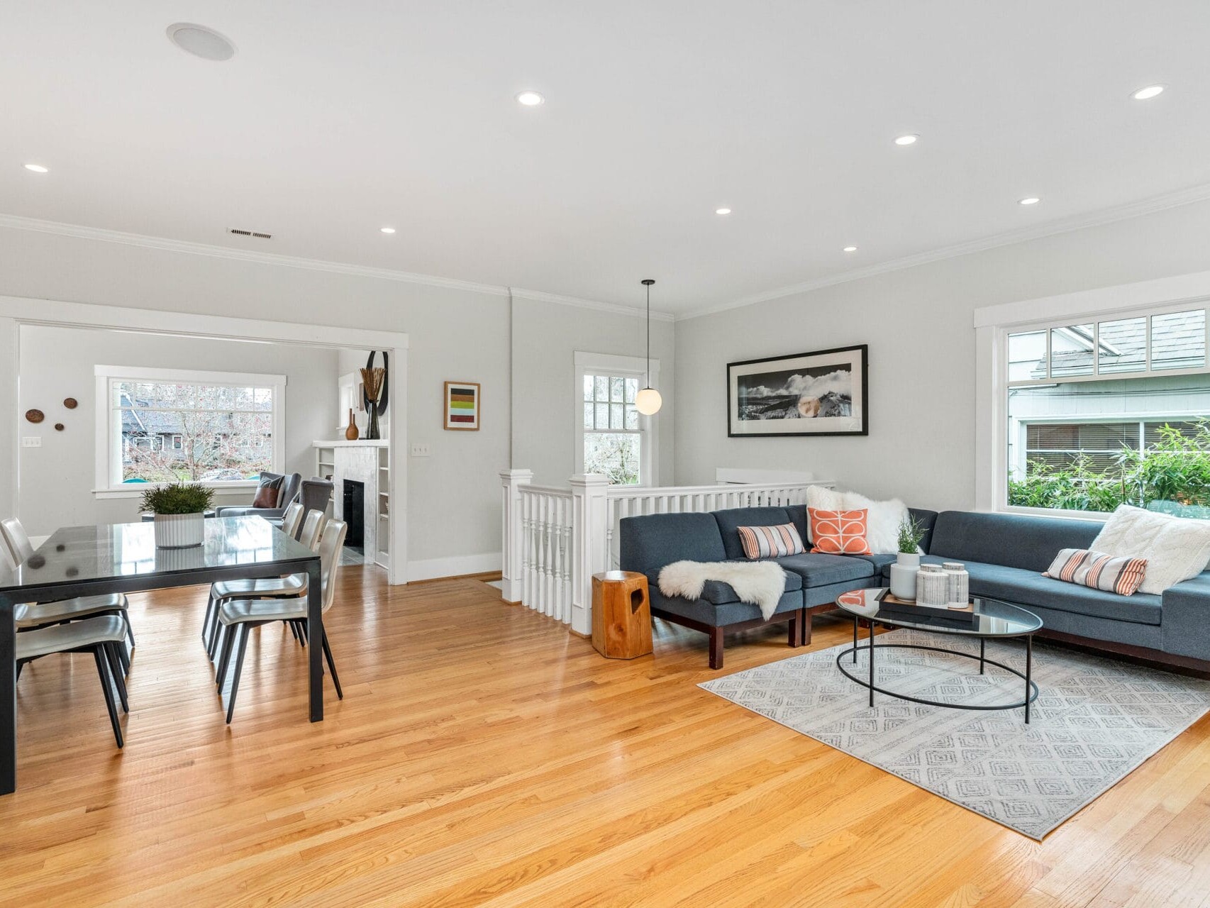 A modern, open-concept living and dining area with hardwood floors. The space includes a gray sectional sofa, a patterned rug, a sleek dining table with chairs, and large windows providing natural light. Minimalist decor adds to the contemporary feel.