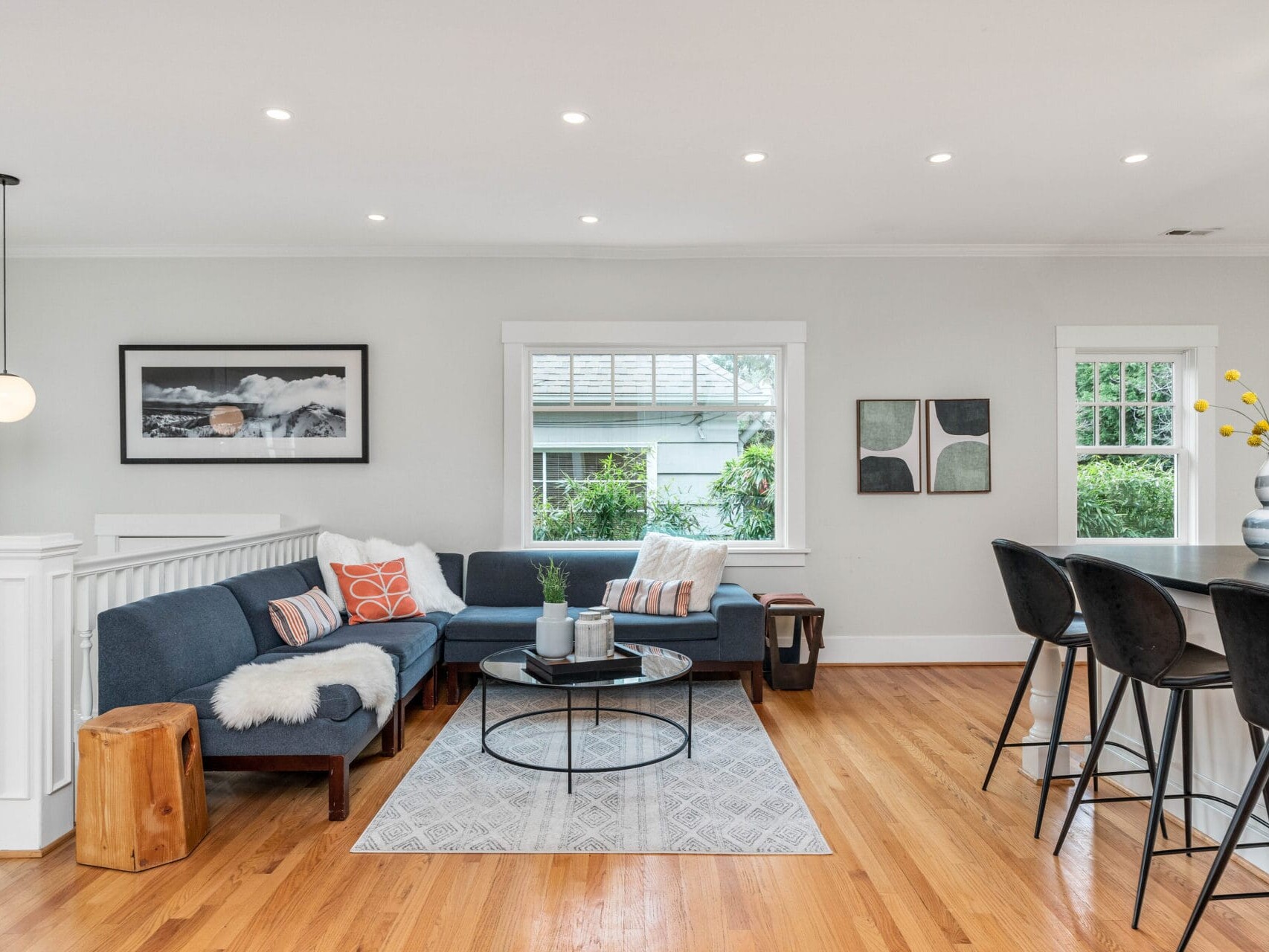 A modern living room with wooden floors, a dark blue sectional sofa, colorful pillows, and a round coffee table on a patterned rug. Art and decor adorn the walls, and a bar with stools is visible. Large windows offer natural light.
