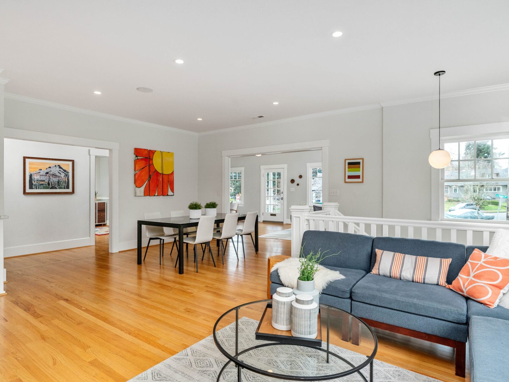 A bright, open-concept living and dining area with hardwood floors. A gray sectional sofa with colorful cushions, a round coffee table, and a dining table with chairs. White walls, recessed lighting, and large windows create a welcoming atmosphere.