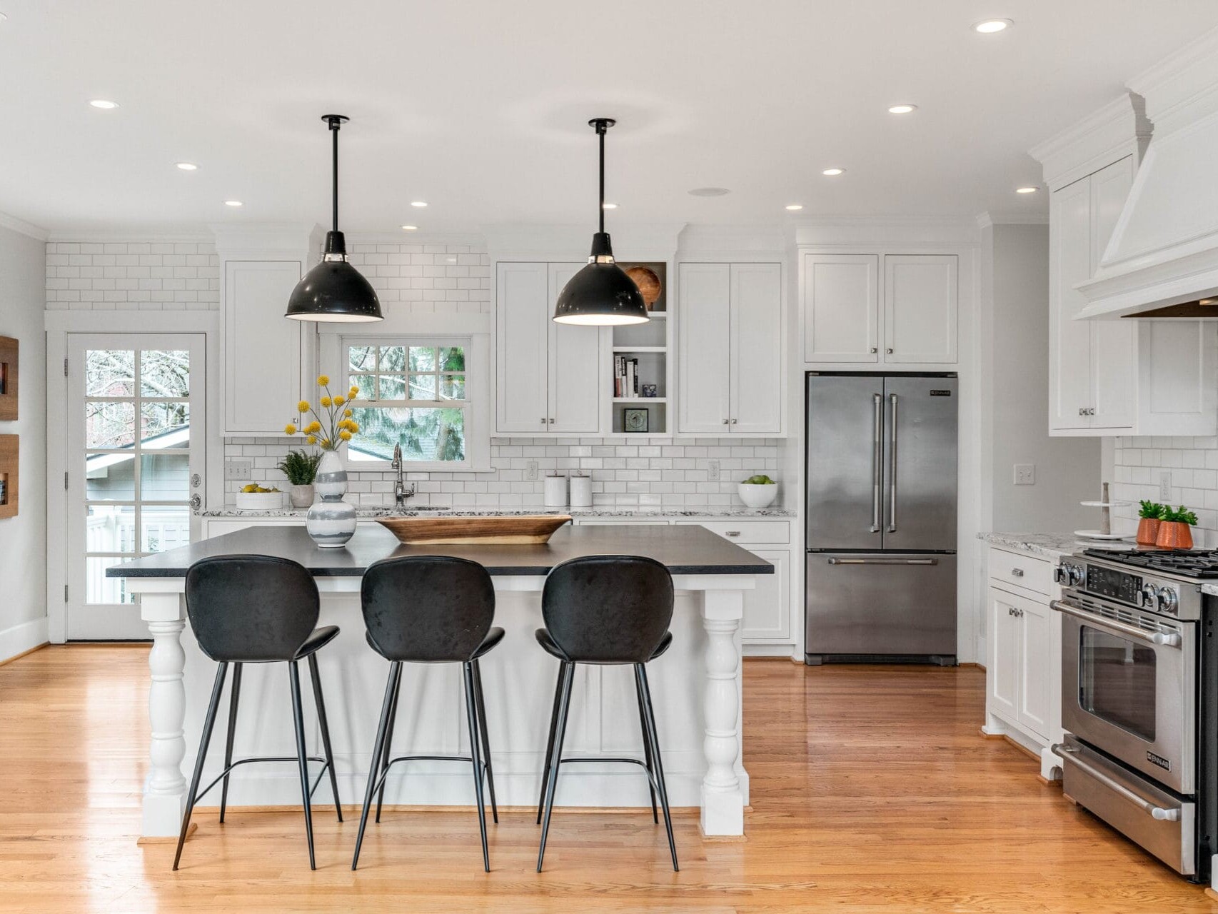 A bright, modern kitchen with white cabinets and subway tile backsplash. A large island features three black barstools and two pendant lights overhead. Stainless steel appliances and wooden flooring complete the space.