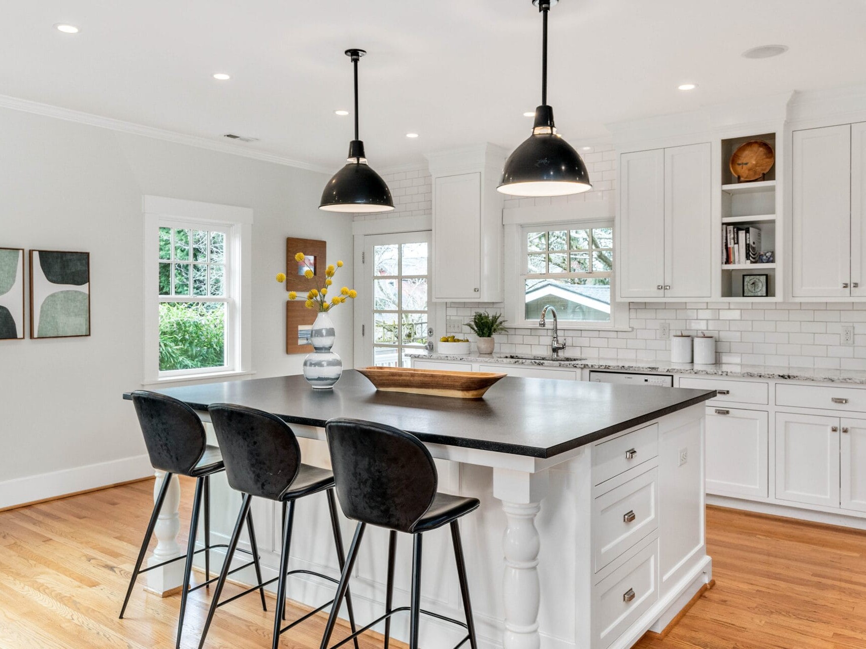 A modern kitchen with a large island featuring three black chairs. White cabinets, a subway tile backsplash, and two black pendant lights are visible. A vase with yellow flowers sits on the island. Wooden floors and abstract wall art complete the scene.