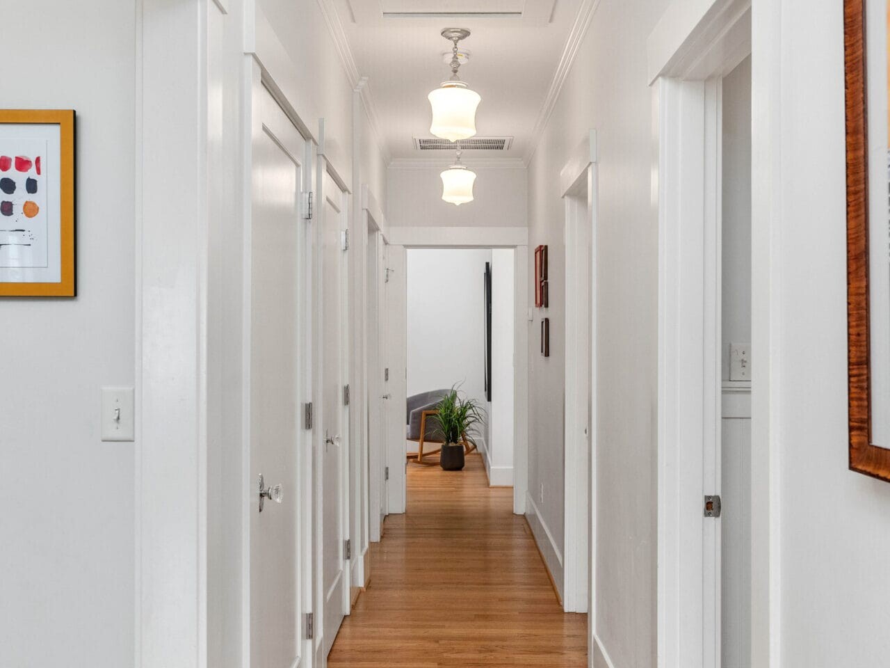 A brightly lit interior hallway with wooden floors and white walls. Several doors and modern light fixtures are visible along the hallway leading to a room with a chair and a potted plant. A framed picture hangs on the right wall.