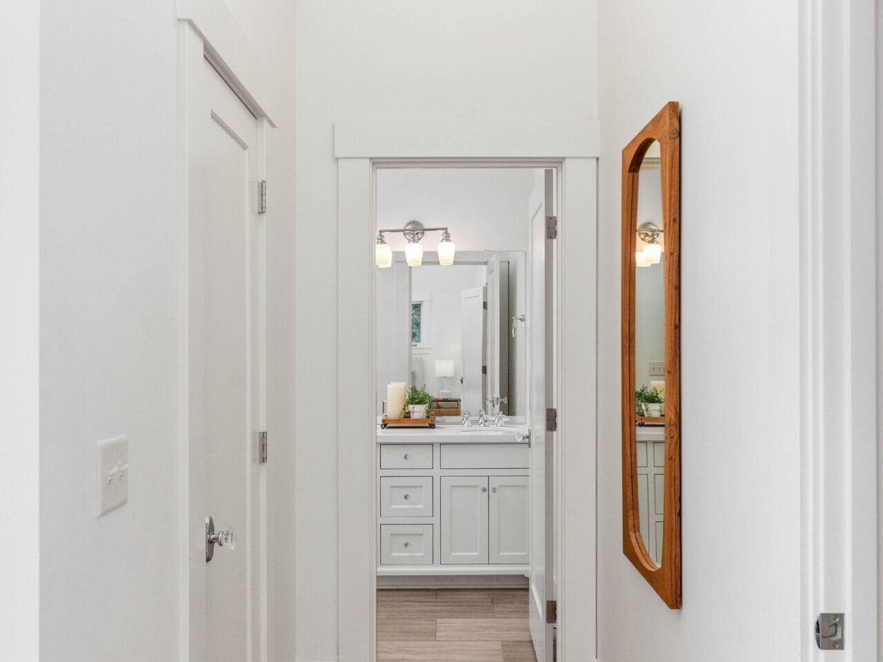 A narrow hallway with a wooden floor leads to a bathroom. There is a wooden framed mirror on the right wall and white doors on both sides. The bathroom at the end has a vanity with a sink, a large mirror, and overhead lighting.