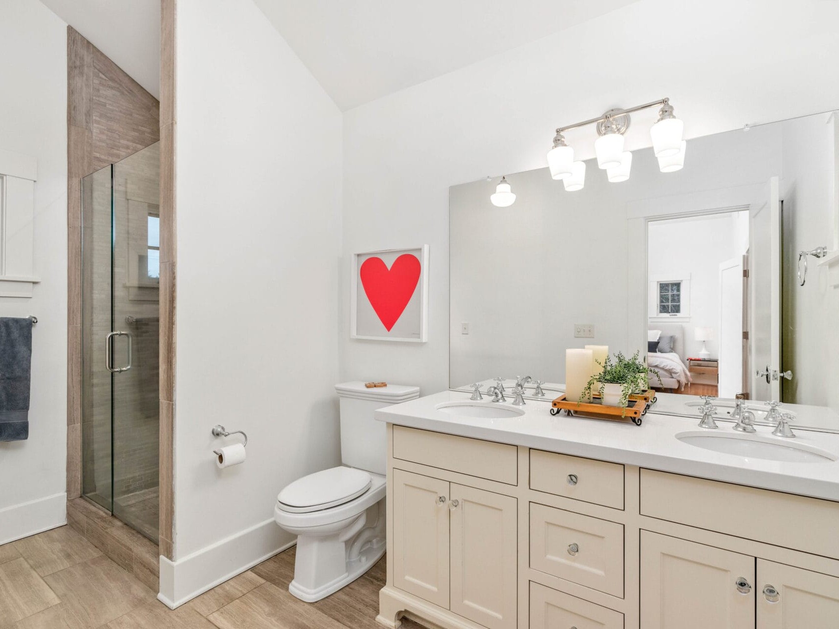 Bright bathroom featuring a double vanity with a large mirror, white cabinets, and a tray with decor. A shower with glass doors is on the left, and a red heart artwork hangs above the toilet. A doorway leads to a bedroom.