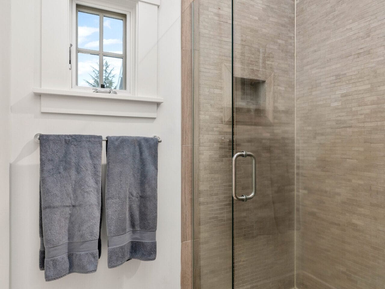 A modern bathroom features a glass-enclosed shower with beige tiles. Two blue towels hang on a towel bar next to a small window showcasing a blue sky with clouds. The floor is tiled in a light wood pattern.