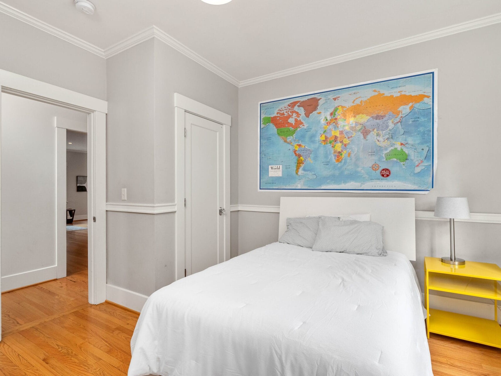 A bedroom with a white bed, a world map on the wall, and a yellow bedside table with a lamp. The room has wooden floors and light gray walls. An open door leads to another room.