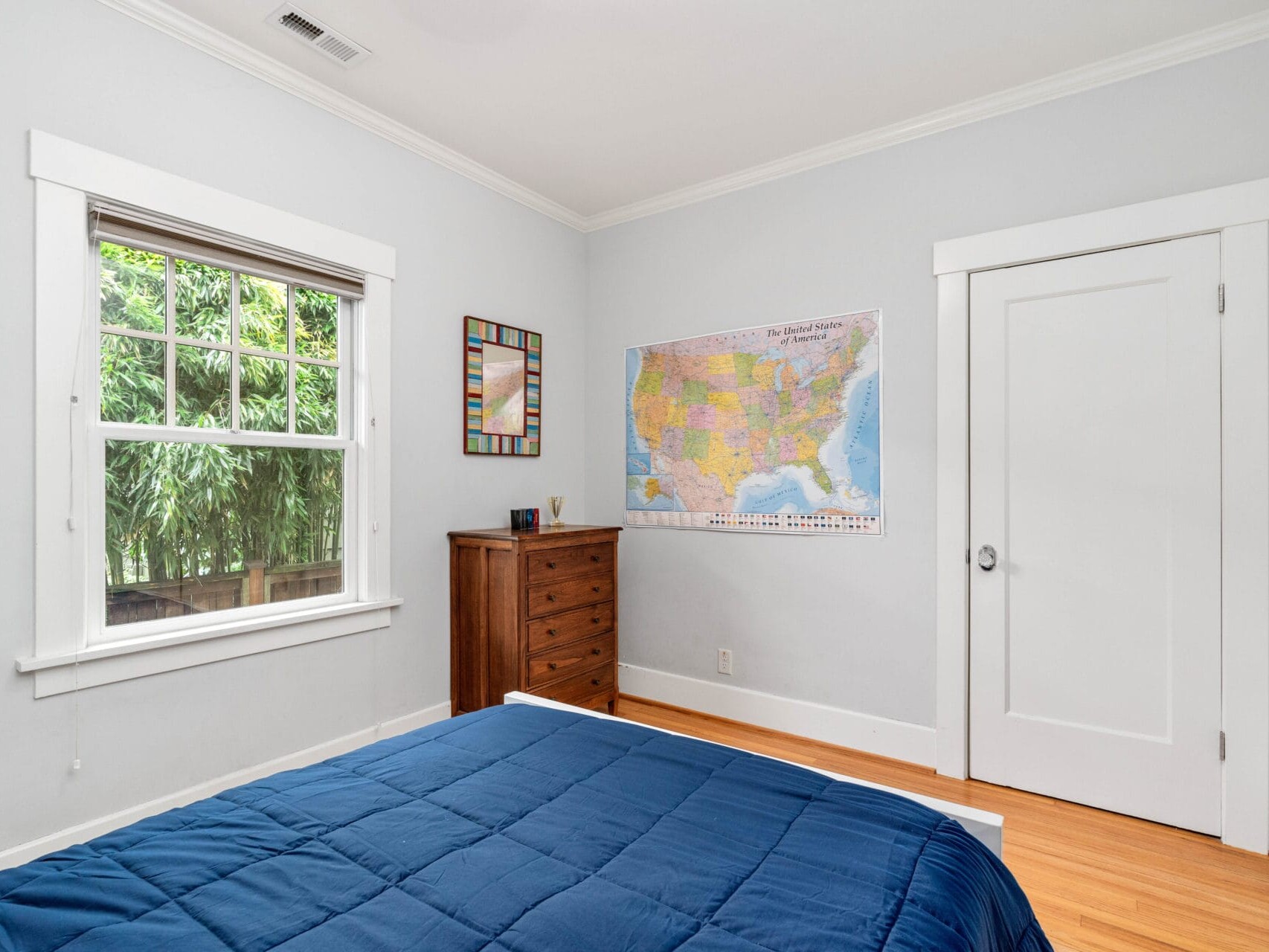 A cozy bedroom with soft gray walls features a large window with greenery outside, a wooden dresser, and a bed with a blue comforter. A colorful map of the United States is displayed on the wall. The room has light wood flooring.