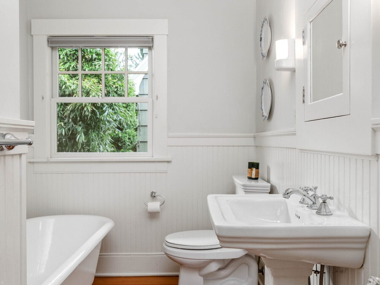 A small, bright bathroom with white walls and a wooden floor. It features a clawfoot bathtub, a pedestal sink, and a toilet. A window with a view of greenery and a wall-mounted cabinet add to the space. Two round mirrors are above the sink.