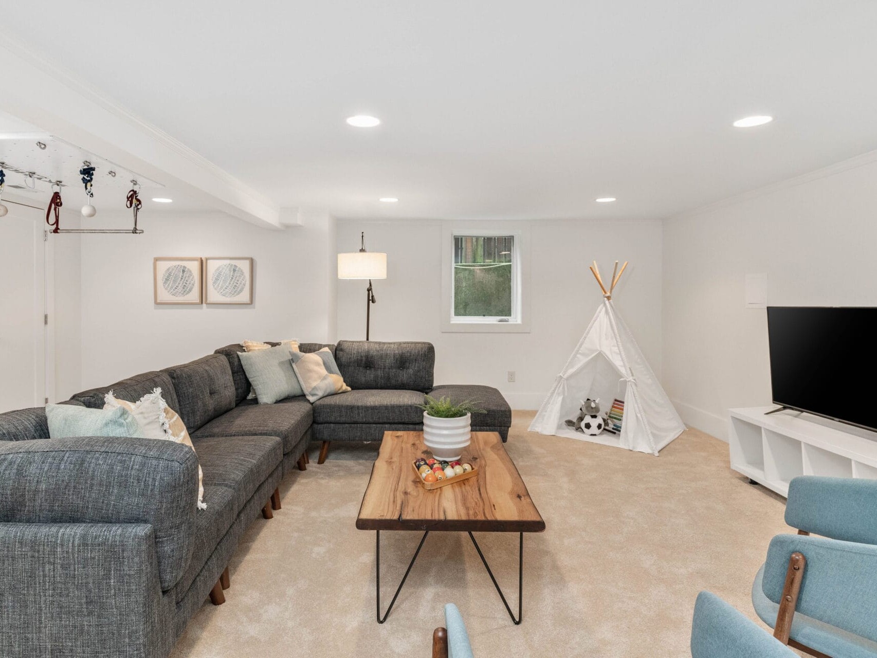 A cozy basement living room with a large gray sectional sofa, a wooden coffee table, and a teepee play tent. The room features a flat-screen TV, framed art on the wall, and soft lighting, creating a comfortable atmosphere.