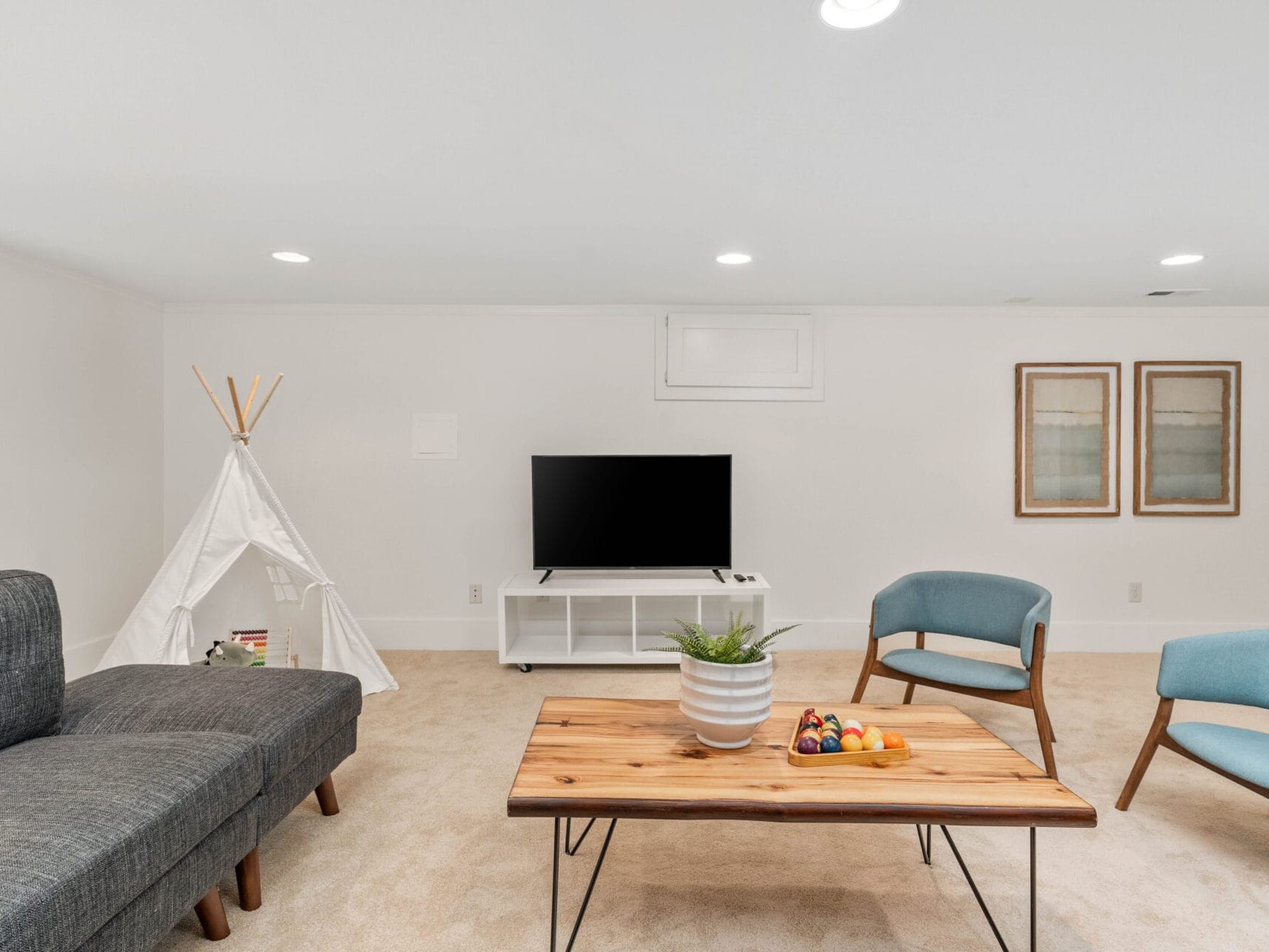 A modern living room with a gray sofa, wooden coffee table, two blue chairs, and a TV on a white stand. A small indoor tent is on the left, and two framed artworks hang on the wall. A bowl of fruit and a potted plant are on the table.
