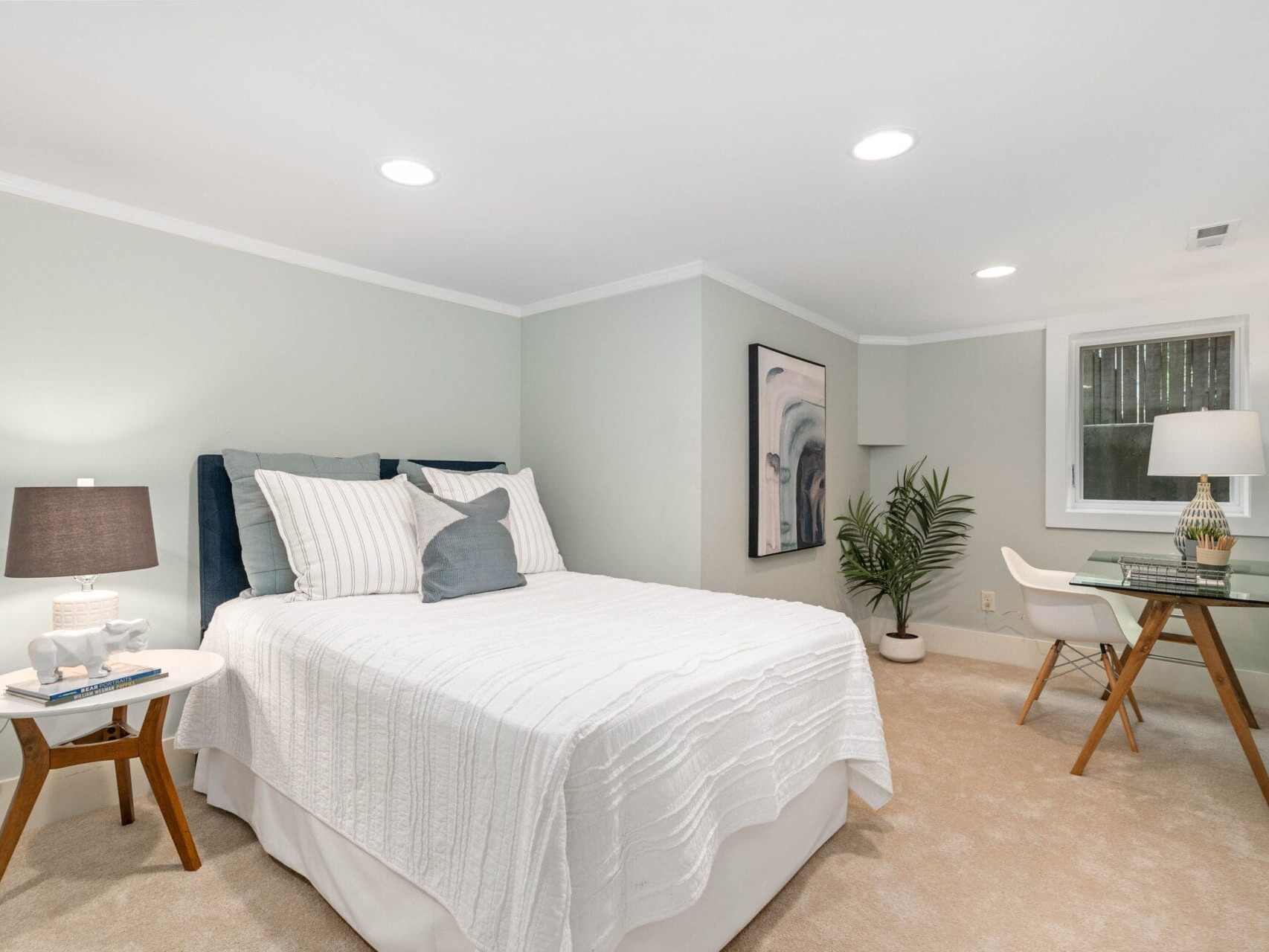 A cozy bedroom with a neatly made bed, side table with a lamp, decorative items, and a small plant. A desk with a chair and lamp is on the right. Soft lighting and abstract wall art enhance the rooms tranquil ambiance.