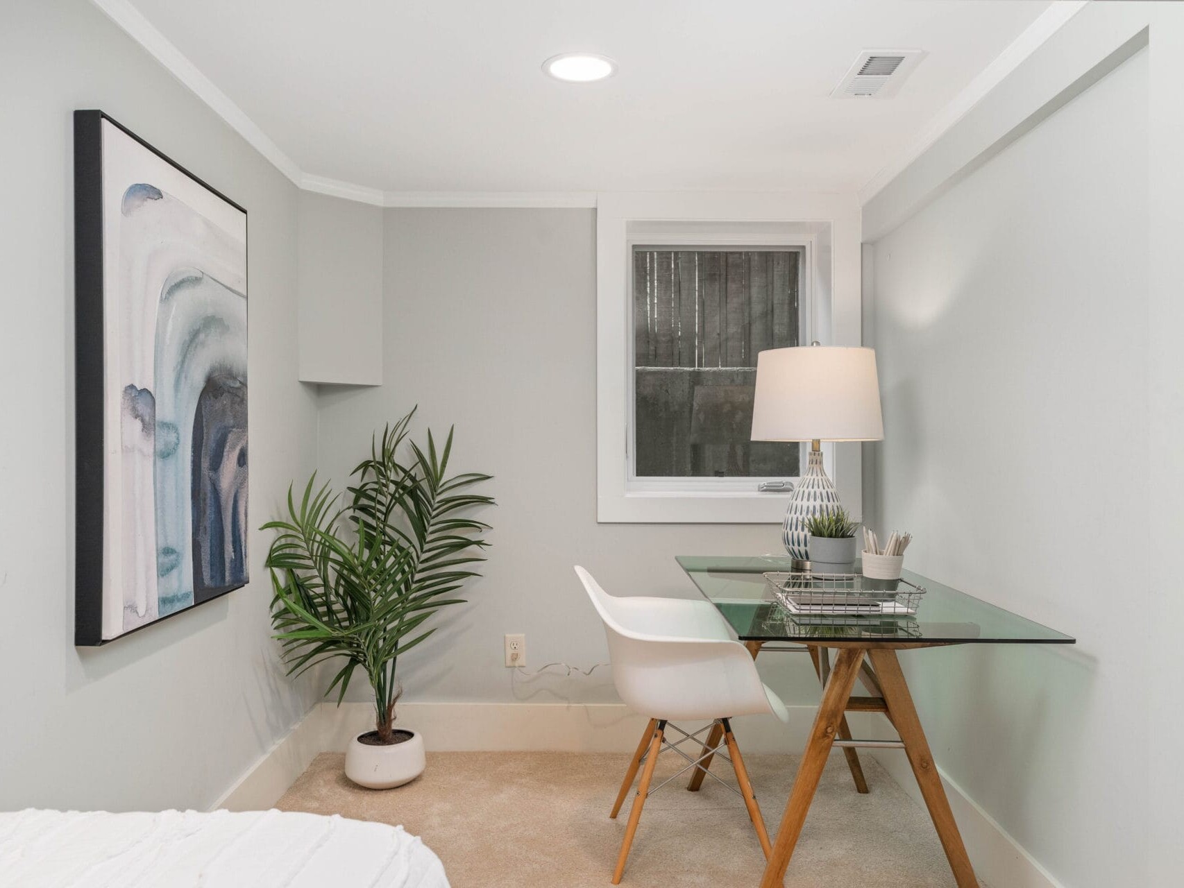 A small, modern home office with a glass desk, white chair, and table lamp. A potted plant is placed on the carpeted floor. Abstract artwork hangs on the light gray wall under a recessed light near a window.