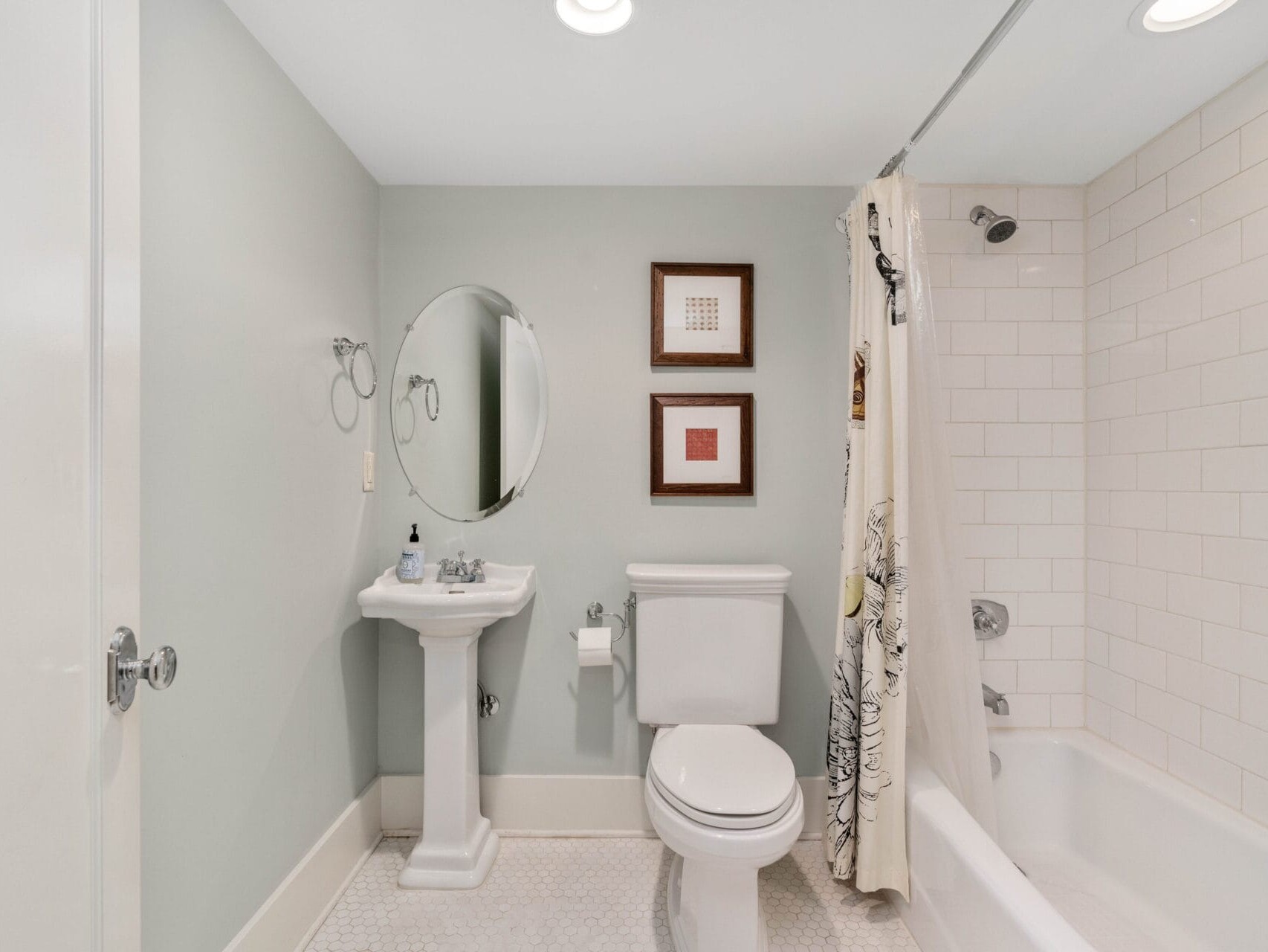 A bathroom with a pedestal sink, oval mirror, and toilet. A bathtub with a shower curtain on the right side, featuring white subway tiles. The walls are painted light gray, and there are two framed pictures above the toilet.